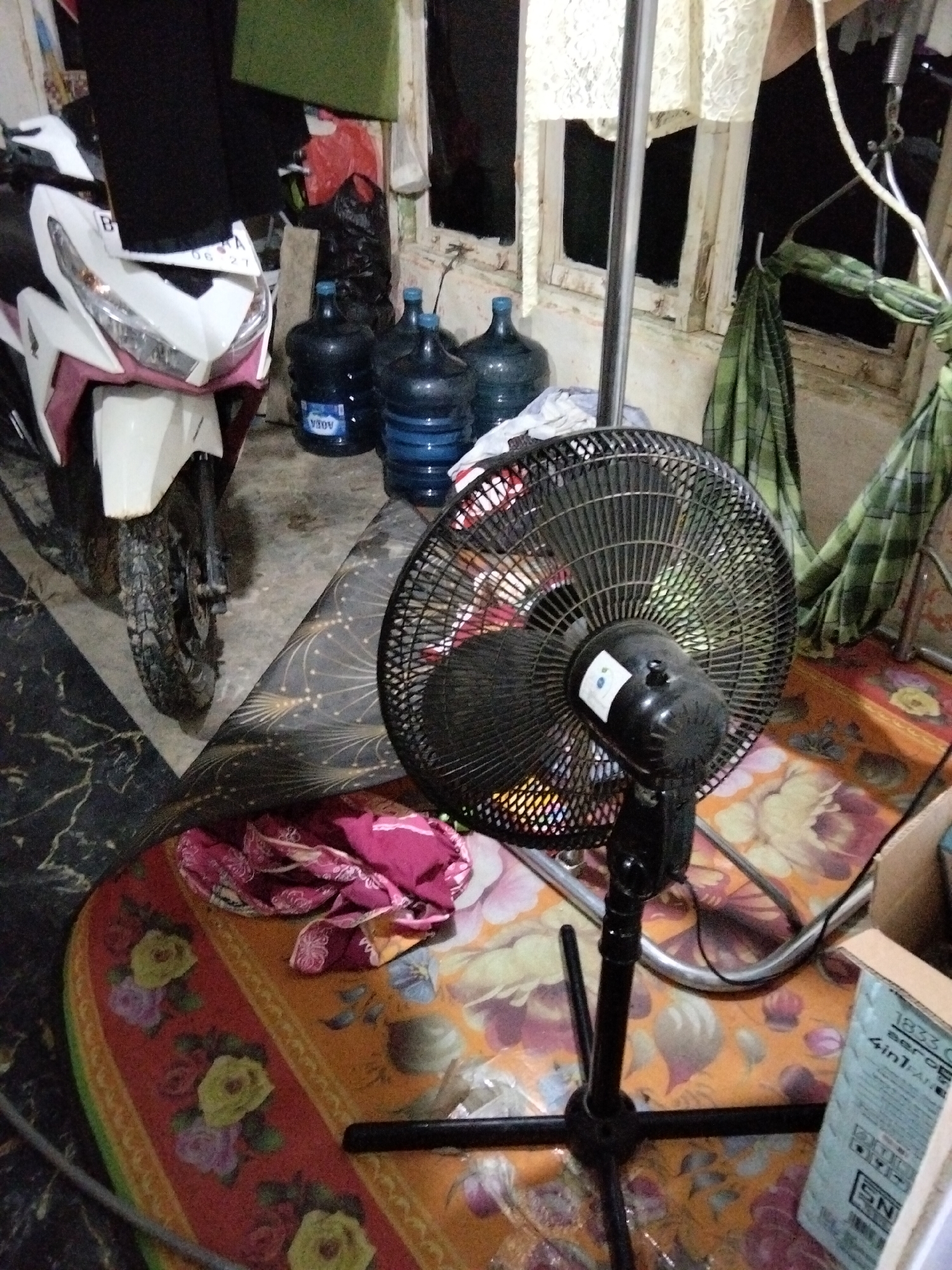 The image depicts an interior scene, likely a room in a residential setting within Benuaraja, Indonesia. In the foreground, a black oscillating fan stands on a colorful, floral-patterned rug, its blades partially visible. To the left, the front portion of a white and pink scooter is visible, parked on a dark, marbled floor. Behind the scooter, several large, blue plastic water jugs are arranged. The background shows a window with a wooden frame, partially obscured by lace curtains and hanging green checkered fabric. The overall impression is one of a somewhat cluttered but functional living space.