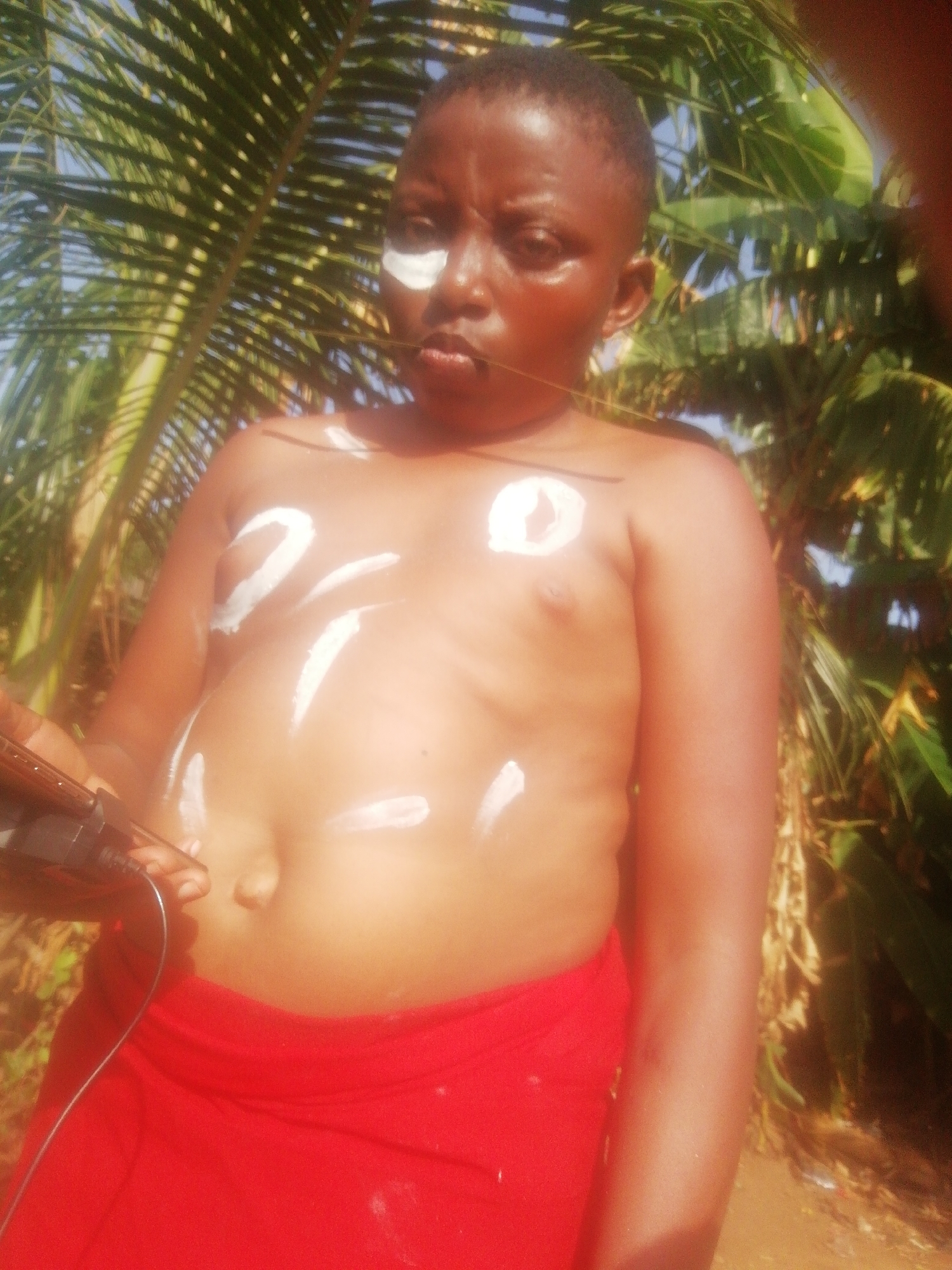 A person stands shirtless in an outdoor location with tropical foliage. The person is wearing a red wrap or cloth around their lower body. There are white markings on their face and upper body. The person is holding an electronic device with a cord in their left hand. The location is Unknown, Unknown.
