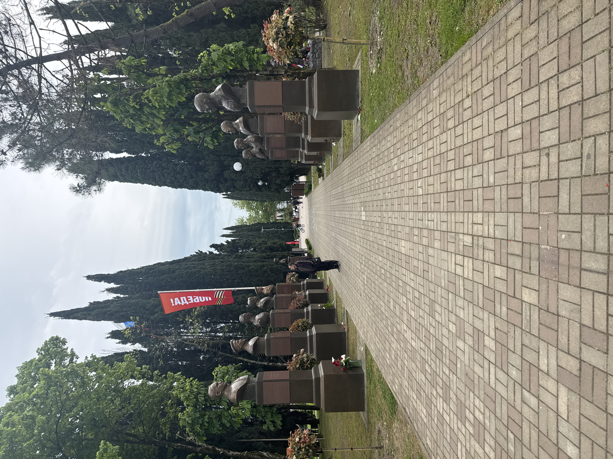 The image shows a memorial site in Sochi, Russia, likely a cemetery or war memorial. A long brick pathway stretches into the distance, flanked by rows of bronze busts mounted on dark-colored pedestals. Some pedestals are adorned with plaques and floral tributes. A red banner with the text "!АДЗЕИАУИ!" and a symbol similar to the ribbon of Saint George flies in the background. Tall, slender trees line both sides of the path, and a person in dark clothing stands on the pathway further away. The sky is overcast.