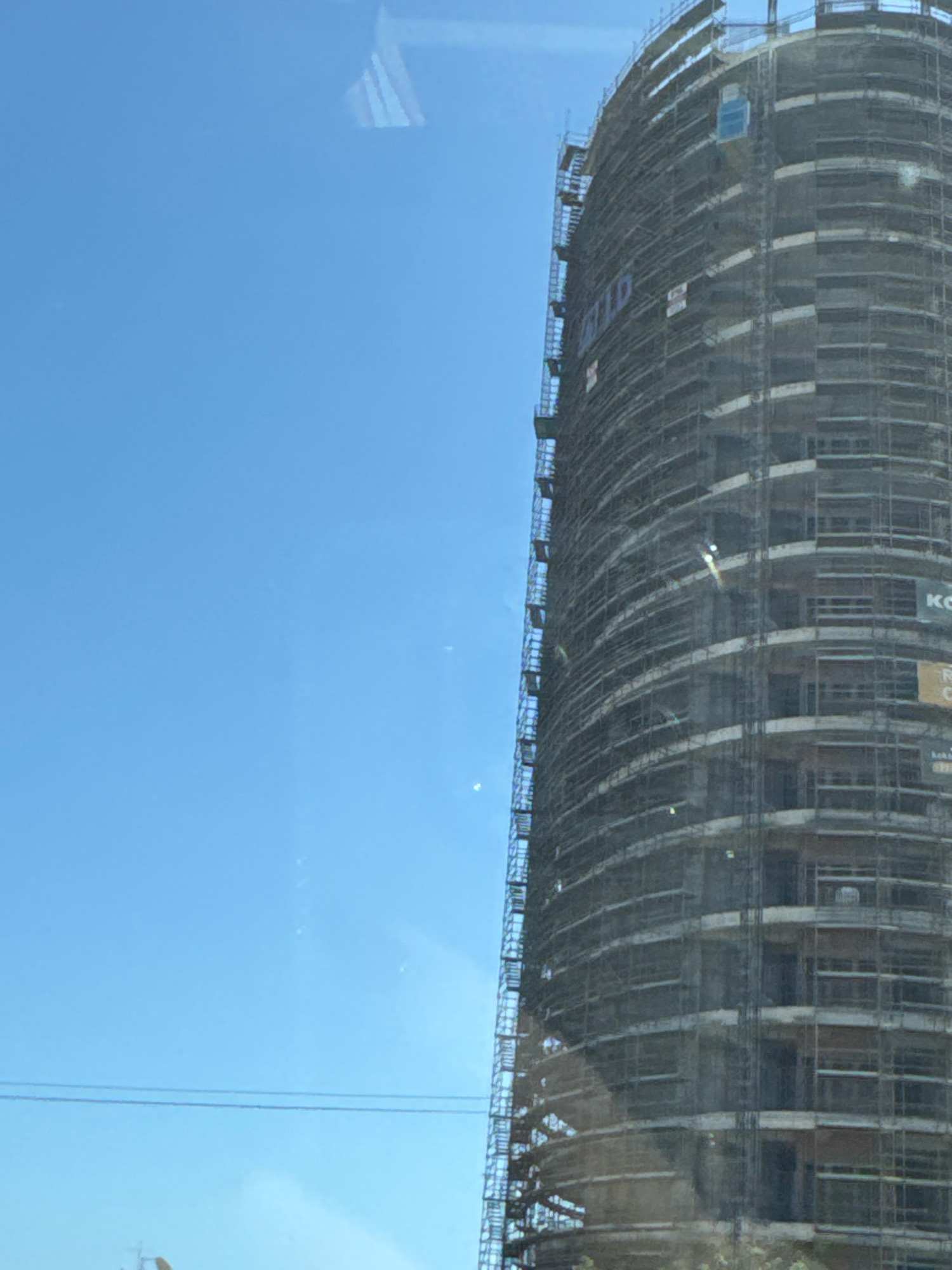 A tall, cylindrical building under construction is visible against a clear blue sky in Agios Tychona Community, Cyprus. The building is encased in scaffolding and netting, with multiple floors and windows in various stages of completion. Several signs are attached to the scaffolding, including one that appears to read "KINGDOM" and another with the letters "KO". Power lines stretch horizontally across the lower portion of the sky. The image seems to be taken from a slightly elevated perspective, possibly from a moving vehicle.