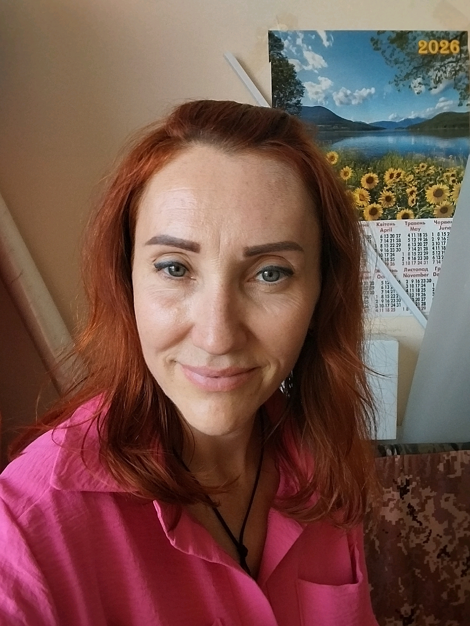 A selfie shows a woman with auburn hair, blue eyes, and light skin. She is wearing a bright pink collared shirt. She is smiling slightly and looking towards the camera. Behind her, on the wall, is a calendar with a scenic image of a lake, mountains, and sunflowers. The calendar is for the year 2026. To the left of the woman, a part of a light-colored wall is visible. To the right of the woman, a camouflage-patterned fabric is visible. The lighting suggests an indoor setting, possibly an office or a home. There are no distinct landmarks visible that would indicate a specific location within the city of Dnipro, Ukraine.