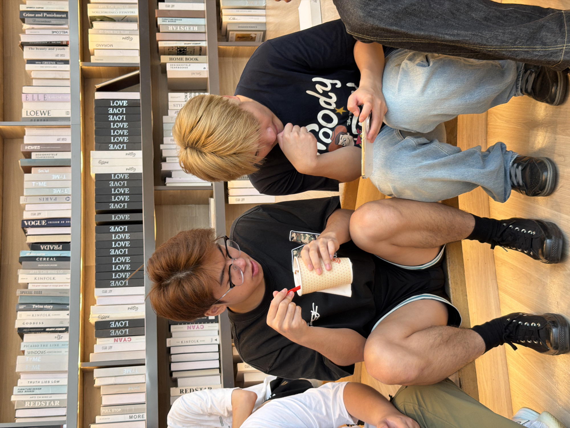 The image depicts two young males seated on wooden steps in front of bookshelves filled with books. The male on the left, with brown hair, wears glasses and a black t-shirt, holding a paper cup with a red straw and a drink. The male on the right, with blonde hair, wears a black t-shirt with a "Woody" graphic and light blue jeans, holding a smartphone. The background shows multiple shelves stocked with books, indicating an indoor environment like a library or bookstore. The lighting appears to be natural, coming from the front. There are no clear indications of the specific location within Liangjiang, China.
