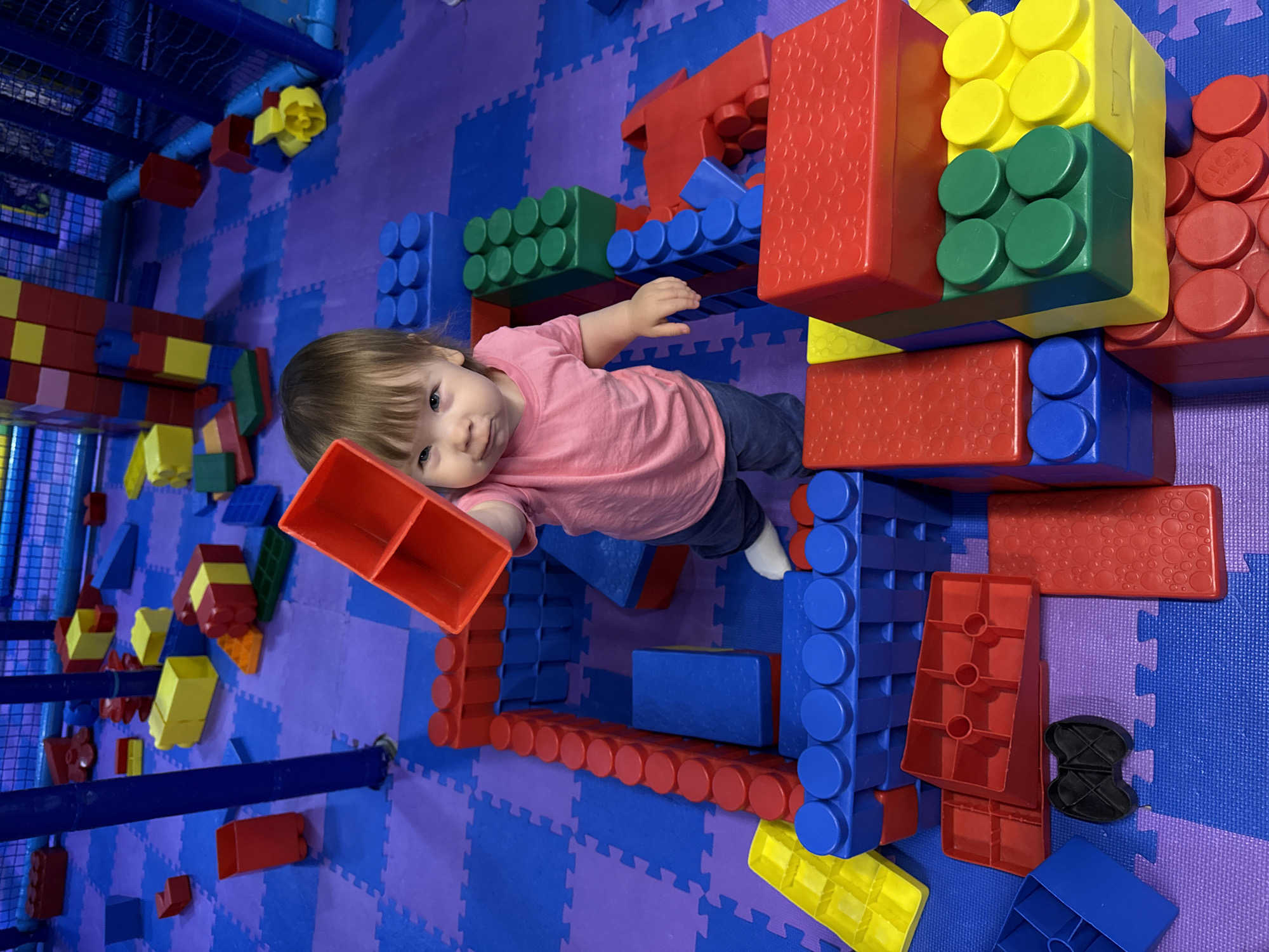 A high-angle shot depicts a toddler playing with oversized building blocks in an indoor playspace. The space features a purple and blue jigsaw-patterned floor. The child, wearing a pink shirt and dark pants, is surrounded by large plastic blocks in red, blue, green, and yellow. The child is holding a red block. Other blocks are arranged to form a low structure around the child. The background shows additional play structures, including netting and a padded climbing area. The location is not specified, but the instruction includes “Перовское сельское поселение, Ukraine,” which is the location.