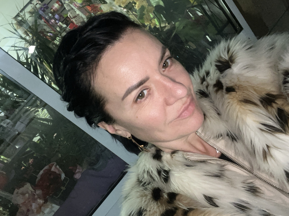 A woman is depicted in a close-up, angled self-portrait, facing slightly towards the viewer with a subtle smile. She has dark hair, styled partially pulled back with some braiding visible on her left side (viewer's right), and brown eyes. Her skin tone is fair. She is wearing a faux fur coat with a cream or off-white base color and dark brown or black leopard-like spots, secured with a visible metallic zipper down the front. A delicate silver-colored chain necklace with a small pendant is visible at her throat. A gold-colored, elongated dangling earring is visible on her left ear (viewer's right). The background consists of a large glass window, showing reflections of what appears to be an interior space. The reflections include green foliage, possibly potted plants or floral arrangements, and various colorful items that resemble merchandise or decorative elements, specifically pink, red, purple, and yellow tones, suggesting a commercial establishment, possibly a florist or gift shop. The window frame is light-colored. The specific location within the city of Smederevska Palanka Municipality, Serbia, cannot be determined from the visual information provided in the image.