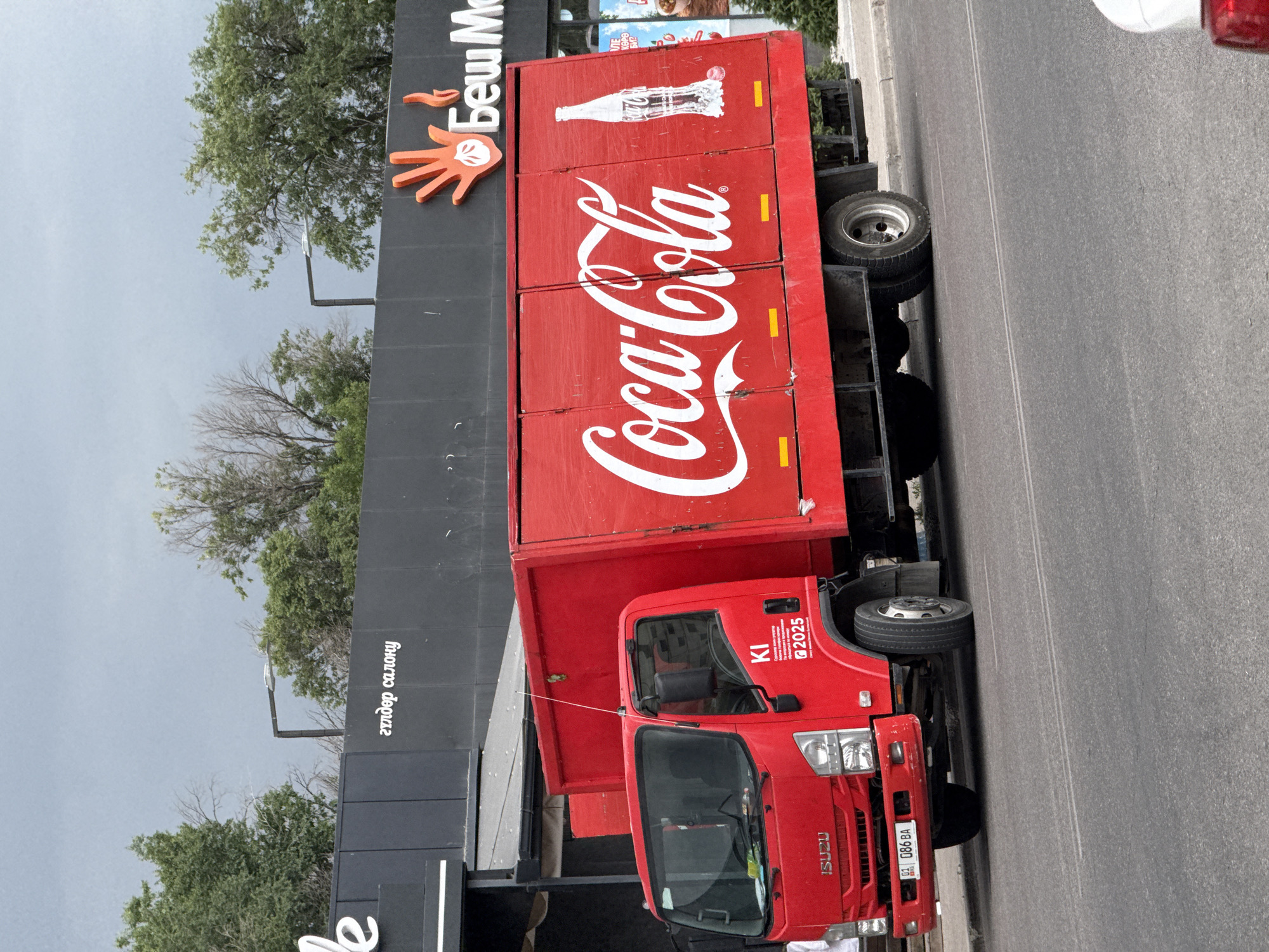 A red Coca-Cola truck is parked on a street in front of a building with a black facade. The truck has the Coca-Cola logo on its side and a picture of a Coca-Cola bottle. On the side of the truck, there is also a white logo with the text "KI" and "2025" beneath it. The building facade has text in a non-English script. The sky is overcast.