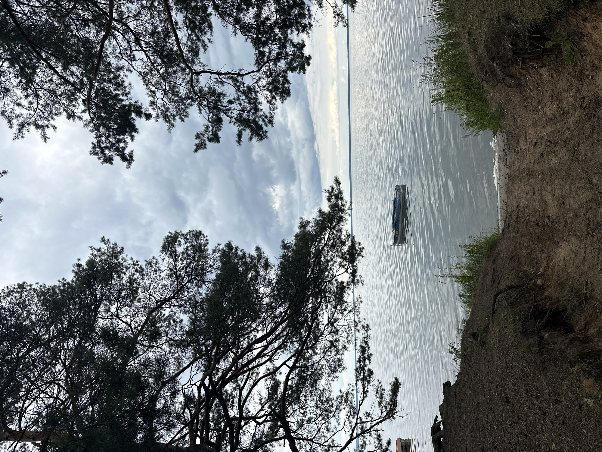 A calm lake reflects a cloudy sky, with pine trees framing the scene from the left and right. In the foreground, a dirt path leads down to the water's edge, where a small, dark boat is moored. Another small boat is partially visible on the left side of the image. The overall atmosphere is serene and natural. The location is the Narach village council, Belarus.