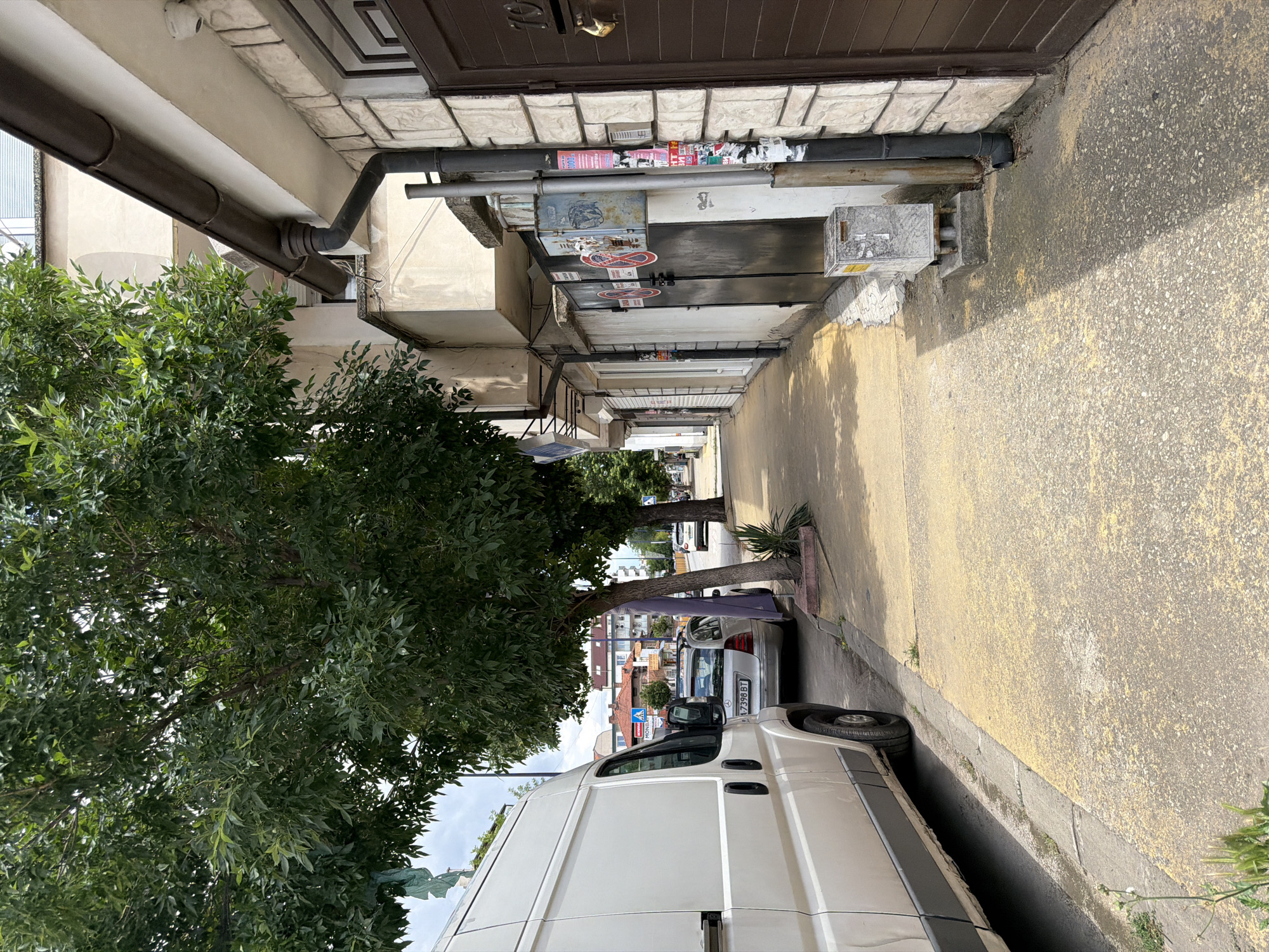 The image shows a street scene in Pazardzhik, Bulgaria. On the left, a white van is parked on the street, facing away from the camera. A sidewalk runs alongside the street, with a yellow-painted surface. Trees line the sidewalk, casting shadows on the ground. To the right of the sidewalk, there is a building with a stone facade. The building has several doors and windows, and a downspout is visible on the side. Advertisements and signs are affixed to the wall and a utility pole. In the background, more buildings and trees are visible, along with a few parked cars. The sky is overcast.
