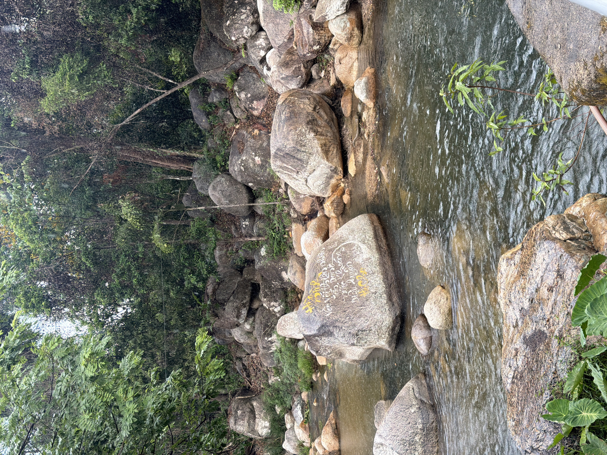 A river in Khanh Vinh, Vietnam. Large, grey rocks line the riverbank and are visible within the water. Dense greenery, including trees and shrubs, grows along the river's edge and among the rocks. The river water is slightly murky, and the current appears gentle. Some of the rocks have yellow writing on them.