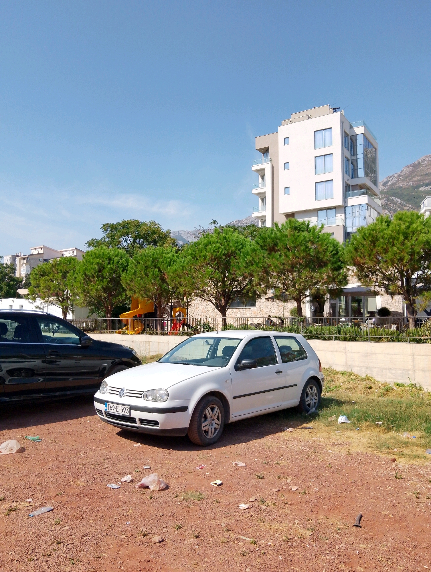This image shows a street scene in Sutomore, Montenegro. In the foreground, a white Volkswagen Golf hatchback is parked on a gravel and dirt surface littered with trash, including plastic bags and bottles. To the left of the white car is a black car. Behind the cars, a low concrete wall separates the parking area from a grassy area with some trees. Further back, a playground with a yellow slide is visible, surrounded by lush green trees. In the background, a modern, multi-story building with white and glass facades stands tall against a clear blue sky. Beyond the building, a mountain range is visible, partially obscured by trees and haze. The overall scene depicts a public space with elements of urban infrastructure, recreation, and nature.