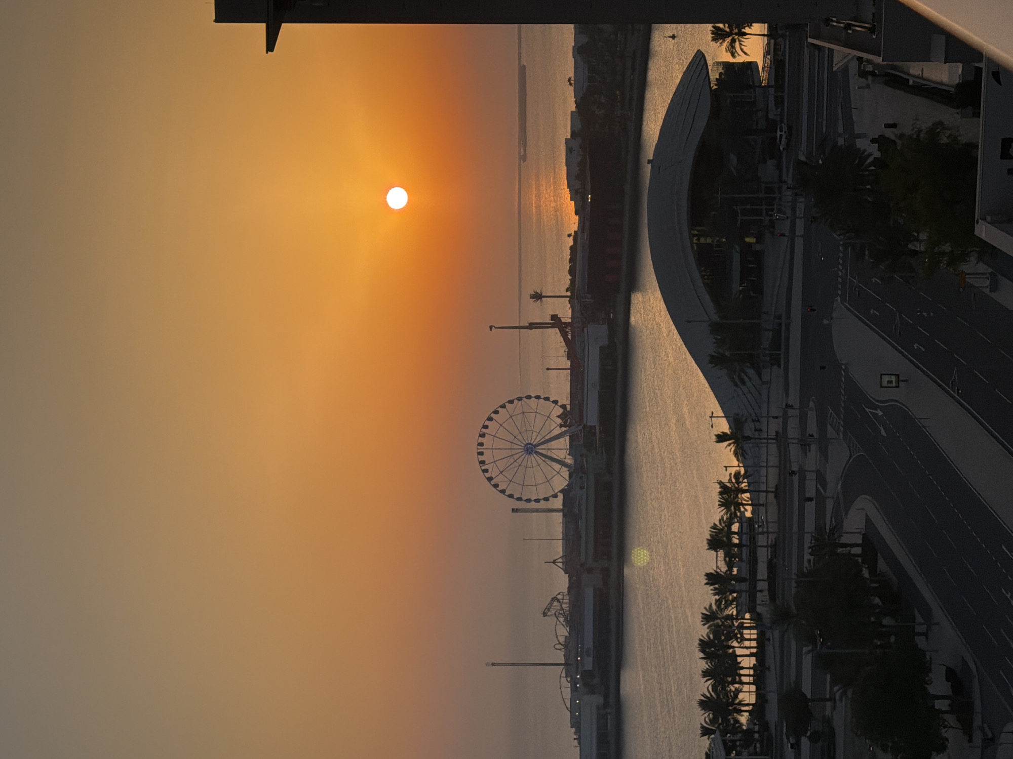 An aerial, high-angle, vertical, medium shot captures a city skyline at sunset. The sun, a bright white circle, is positioned in the upper right quadrant of the frame, casting a warm orange glow across the sky and reflecting on the calm ocean. In the distance, a large Ferris wheel dominates the skyline, alongside other amusement park rides and buildings. The water stretches to the horizon, where a distant ship is visible. In the foreground, a modern, curved building with a large, arched roof is situated near a road lined with palm trees. The road appears empty, with only a few distant vehicles visible. The overall scene is serene and bathed in the golden light of sunset.