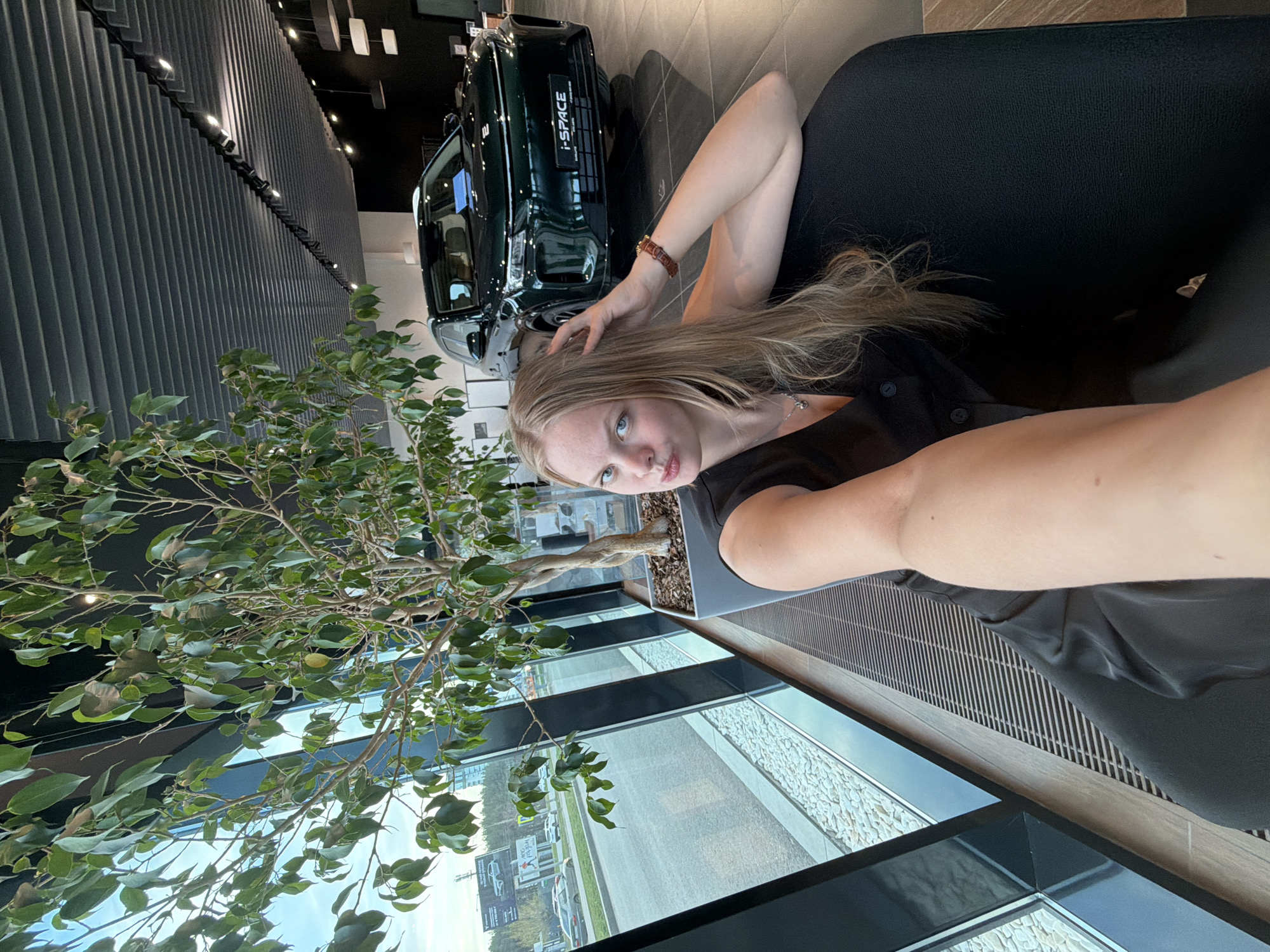 A woman is taking a selfie indoors, with a green car, possibly a Lynk & Co 01 SUV based on the I-Space plate, visible behind her. She is positioned near a large window with a tree in a planter box by the window. Outside the window, a view of a city and a sign with a logo, potentially "ТИТАН СТРОЙ", can be seen. Based on the sign and the architecture, the location is possibly in Izhevsk, Russia. The woman has long blonde hair and is wearing a black top. She has one arm raised to her hair. She is partially sitting on black furniture. The building has modern architecture, and the lighting is bright.