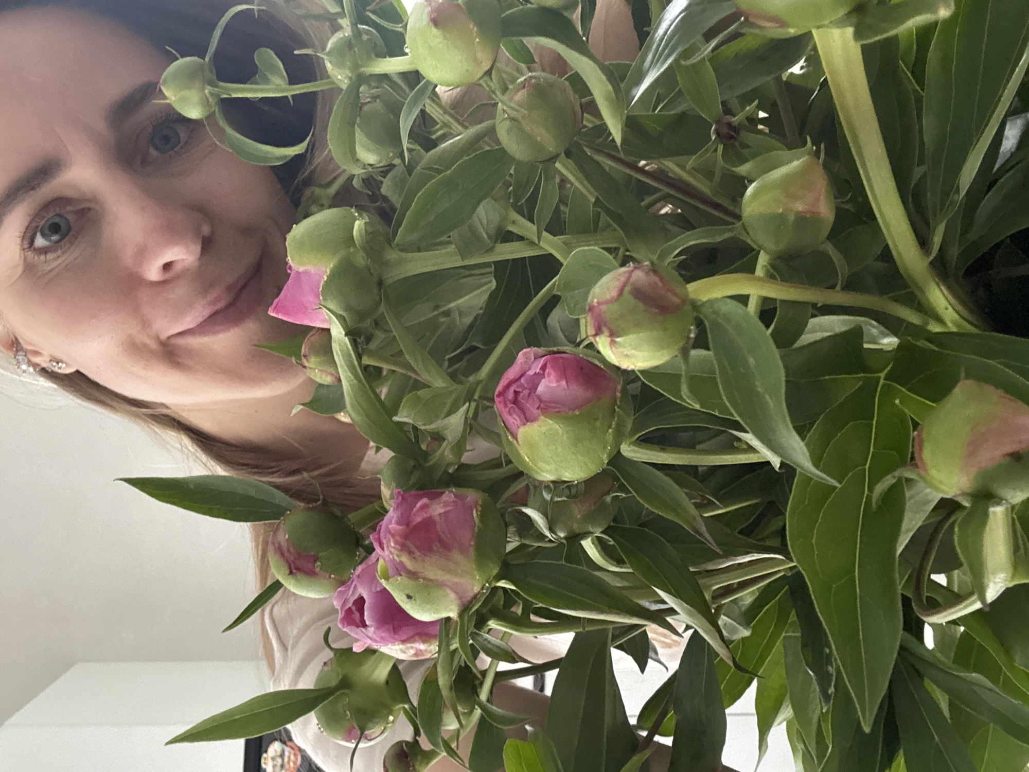 A medium shot captures a woman's face partially visible on the left side of the frame, looking towards the viewer with a gentle, subtle smile. Her light blue eyes are prominent. She is surrounded by a lush bouquet of peonies that fills the center and right portions of the image. The peonies are mostly still in bud form, showcasing vibrant green sepals with hints of pink petals beginning to emerge or partially opened. Many buds display distinct pink coloration at their tips or where they are starting to unfurl. The bouquet consists of numerous green leaves and stems, creating a dense floral arrangement. Part of the woman's light brown hair and a small, sparkly earring are visible on the lower left. The background appears to be a plain, light-colored indoor surface, likely a wall or ceiling, suggesting bright, ambient lighting. No text is visible in the image. The scene takes place in Chaikovsky, Russia.