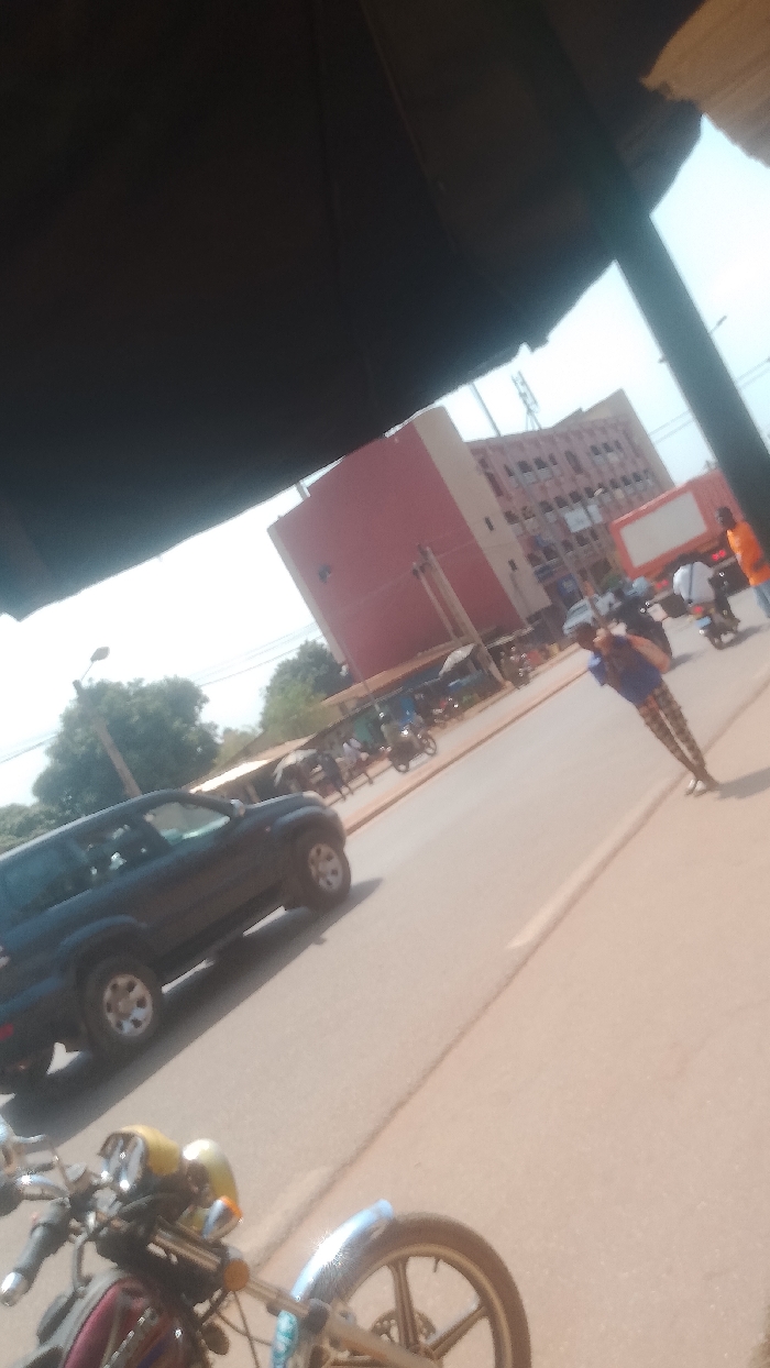 The image displays a street scene in Korhogo, Côte d'Ivoire, captured from a shaded vantage point, with a dark overhead structure obscuring the upper left quadrant. The environment is brightly lit.
In the immediate foreground, the handlebars and front wheel of a motorcycle or moped are partially visible at the bottom of the frame. The midground features a light brown asphalt road with a subtle white dividing line. A dark-colored SUV is positioned on the left side of the road, facing away from the viewer. A pedestrian, wearing a blue shirt and patterned trousers, stands on the right side of the road, appearing to observe the surroundings. Multiple individuals on motorcycles/mopeds are visible further down the road. On the left, behind the SUV, a series of open-air stalls or small shops with light-colored awnings line the roadside. Green foliage from trees is visible behind these structures, and a street light pole stands on the left.
The background is dominated by a multi-story building constructed with reddish-brown facades and numerous rectangular windows. To its right, a large, rectangular, white billboard-like structure is present. The sky above is clear.