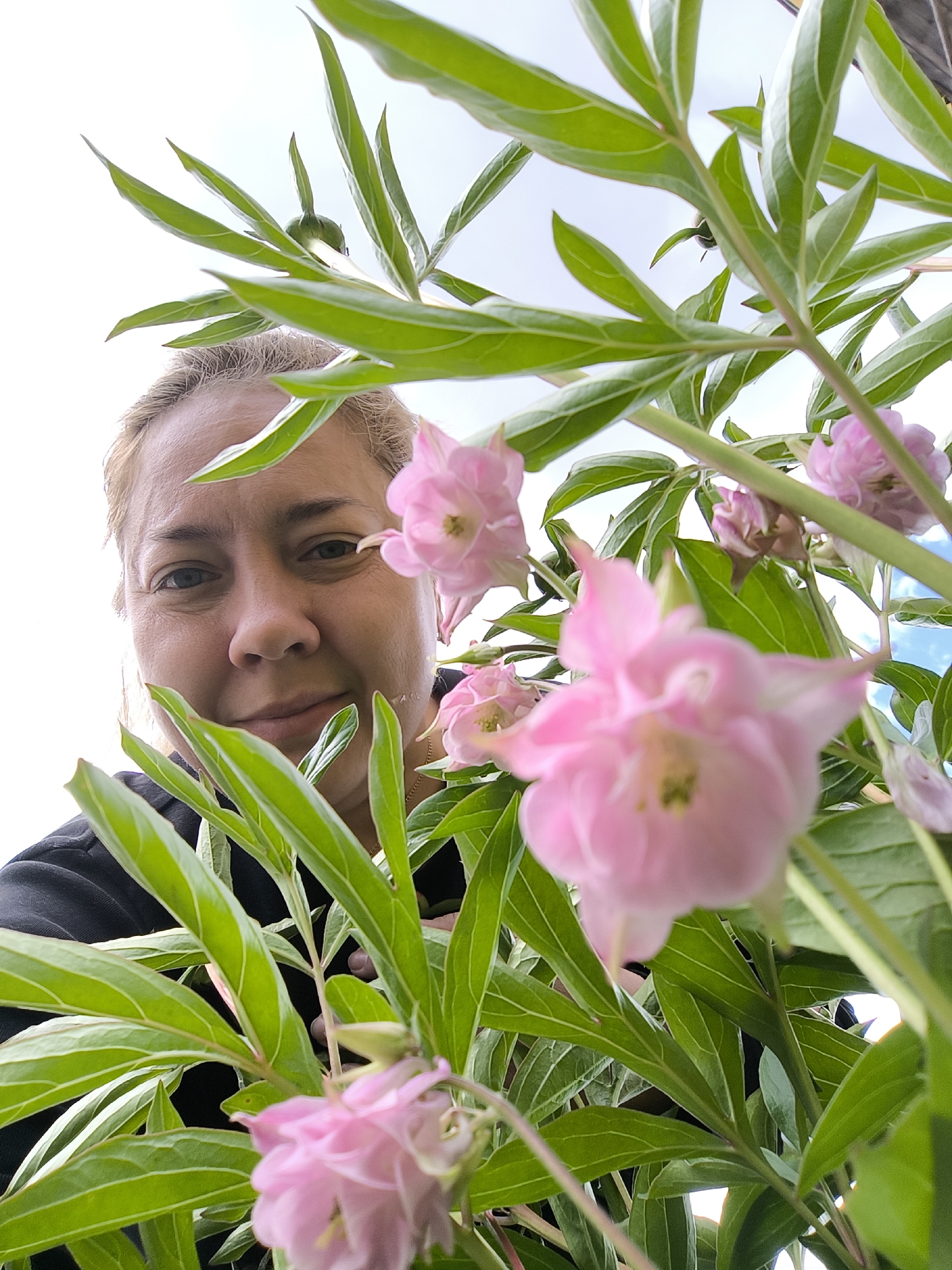 A woman with blond hair and light skin is partially obscured by foliage and pink flowers. She is wearing a black shirt and smiling slightly. The leaves are light green with white edges. The sky is visible in the background. The location is unspecified.