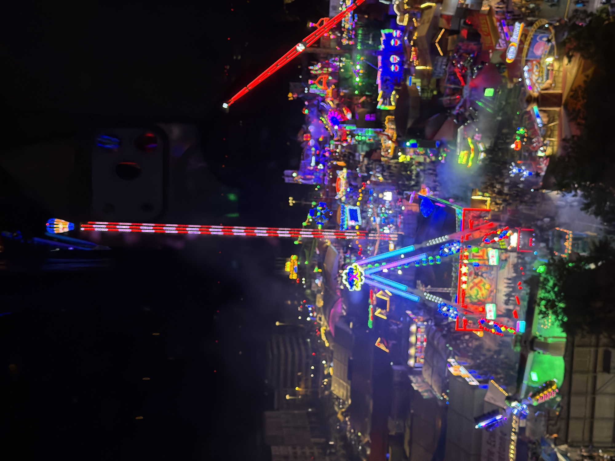 The image depicts a night view of a bustling amusement park in the city of Luxembourg, Luxembourg. The park is illuminated with a vibrant array of colorful lights from various rides and attractions. In the foreground, a tall, red and white striped tower with a yellow and blue top structure is prominent. To the right, a red arm extends upwards, characteristic of a thrill ride. The middle ground features a variety of rides, including a colorful, multi-tiered structure with people on it, a swing ride with bright lights, and other illuminated attractions. The background shows buildings and streetlights, suggesting the park is located within the urban environment of Luxembourg City. Numerous people are visible, milling about and enjoying the attractions, creating a lively atmosphere. Smoke or mist is present in some areas, possibly from fog machines or steam from food vendors. The overall scene captures the energetic and festive ambiance of a night at an amusement park.