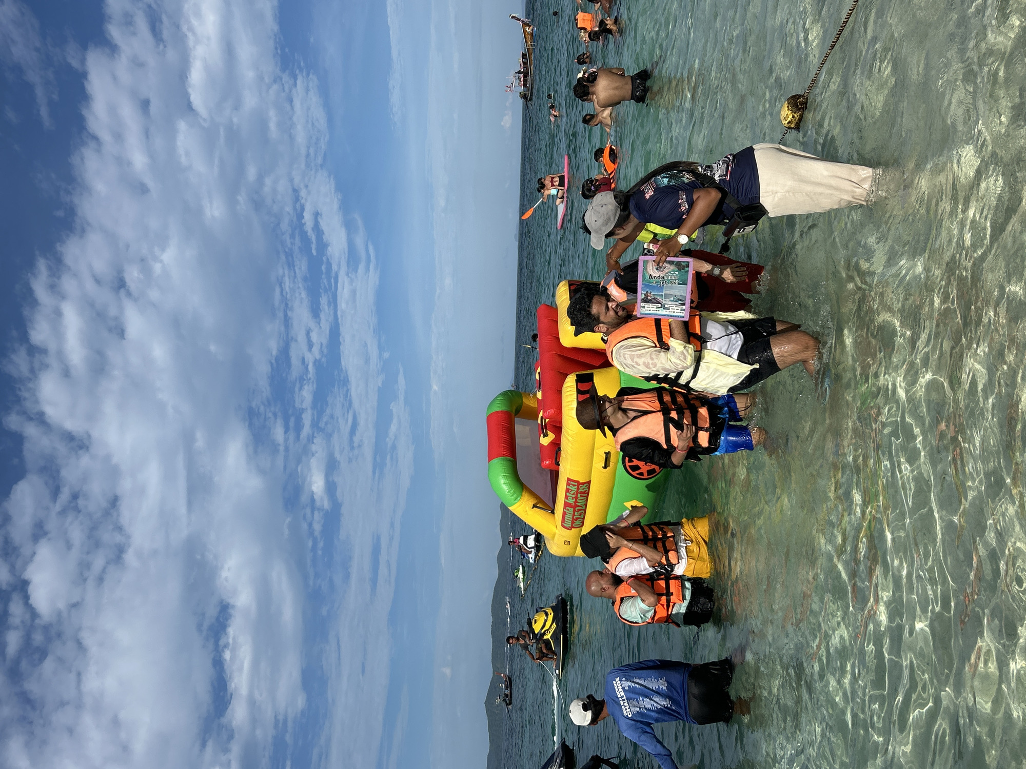 The image depicts a vibrant beach scene in Tambon Phru Nai, Thailand, under a partially cloudy sky. In the foreground, several people are wading in the clear, shallow water, many wearing bright orange life vests. Some are interacting with a large, inflatable water toy that resembles a vehicle, which is yellow, red, and green. To the left, a person in a blue shirt with "2023 WU SCHOOL CHALLENGE" printed on it stands. Further back, various individuals are engaged in water activities, including jet skiing and paddleboarding. Several jet skis are visible, some in motion, and a few boats are dotted on the horizon. The water is remarkably transparent, revealing the sandy bottom and schools of small fish. The overall atmosphere suggests a lively tourist destination where recreational water sports are popular.