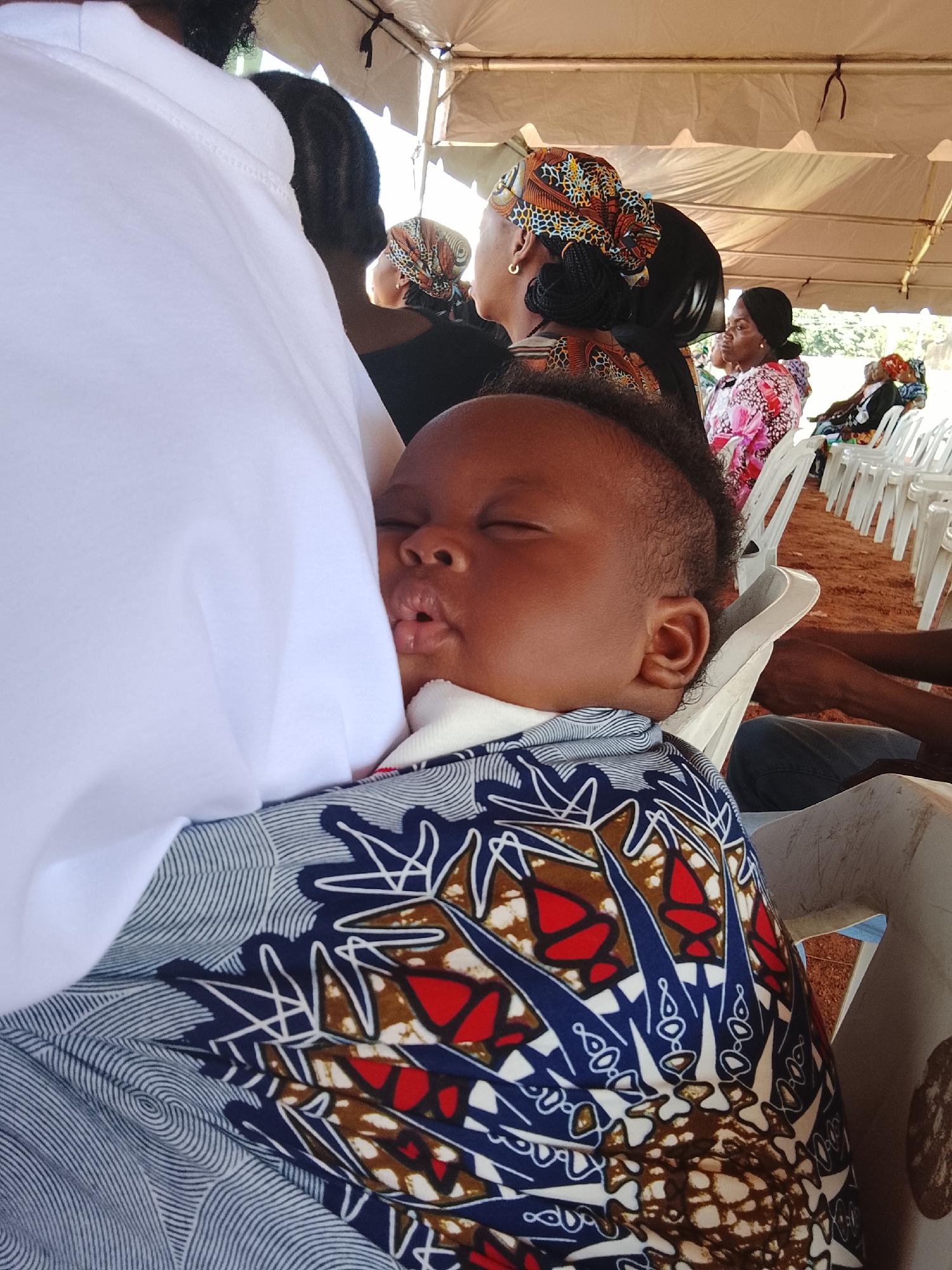 A sleeping baby is held in a fabric wrap in the foreground of the image. The baby is dark-skinned with closed eyes and their mouth is slightly open, with the tongue visible. They are wearing a white garment around their neck and a colorful fabric wrap with a blue, white, and red pattern covering their body. The person holding the baby, who is mostly out of frame on the left, is wearing a white garment.
In the background, several people are seated on white plastic chairs under a white tent. The ground appears to be made of reddish-brown dirt. Some of the individuals in the background are wearing headscarves with vibrant patterns. The scene is set outdoors, under the tent, suggesting a gathering or event. The overall atmosphere appears to be calm and relaxed, with the focus on the sleeping infant. This scene could be taking place at a community event or a family gathering in Akwanga, Nigeria.