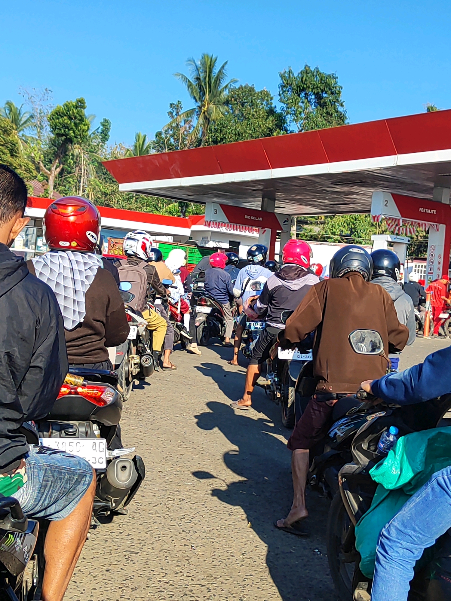 The image depicts a queue of motorcyclists at a gas station, likely in Bondowoso, Indonesia, under a clear blue sky. The gas station features a red and white canopy with signage indicating "BIO SOLAR" and "PERTALITE". Several individuals are seated on their motorcycles, many wearing helmets of various colors, including red, white, and black. Some are also wearing casual clothing like t-shirts, hoodies, and jeans. The foreground shows a person in a black jacket and denim shorts, and another person in a blue jacket. The motorcycles are lined up on an asphalt road. Tropical trees and vegetation are visible in the background, suggesting a warm climate. The overall scene portrays a typical day at a gas station with people waiting to refuel their vehicles.