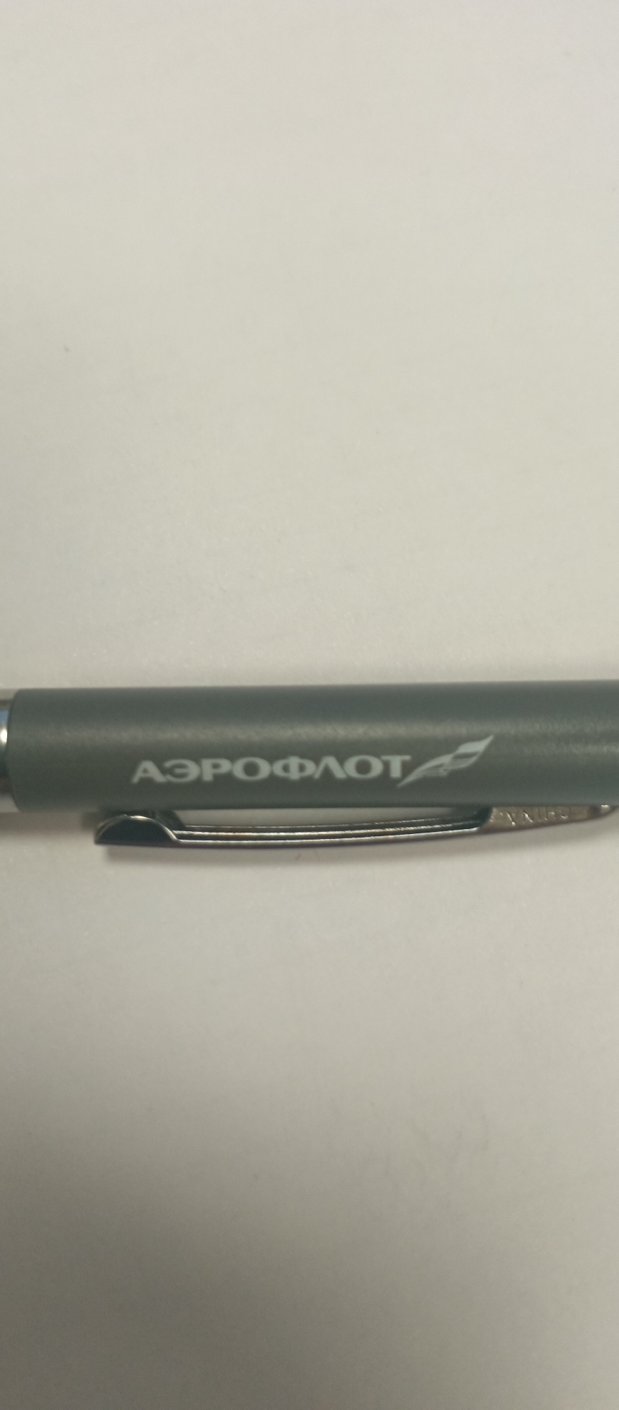 This media file shows a close-up of a grey pen with white text and a logo on it. The pen is lying on a white surface. The text visible on the pen is in Cyrillic script and reads "АЭРОФЛОТ" which is the Russian word for Aeroflot, a well-known Russian airline. To the right of the text is a white logo that appears to be a stylized bird or airplane. Below the text and logo, there is a silver metal clip attached to the pen. The lighting suggests the image was taken indoors under artificial light, and there are no indications of weather or time of day. The focus is solely on the pen, and no people or other objects are visible in the frame.