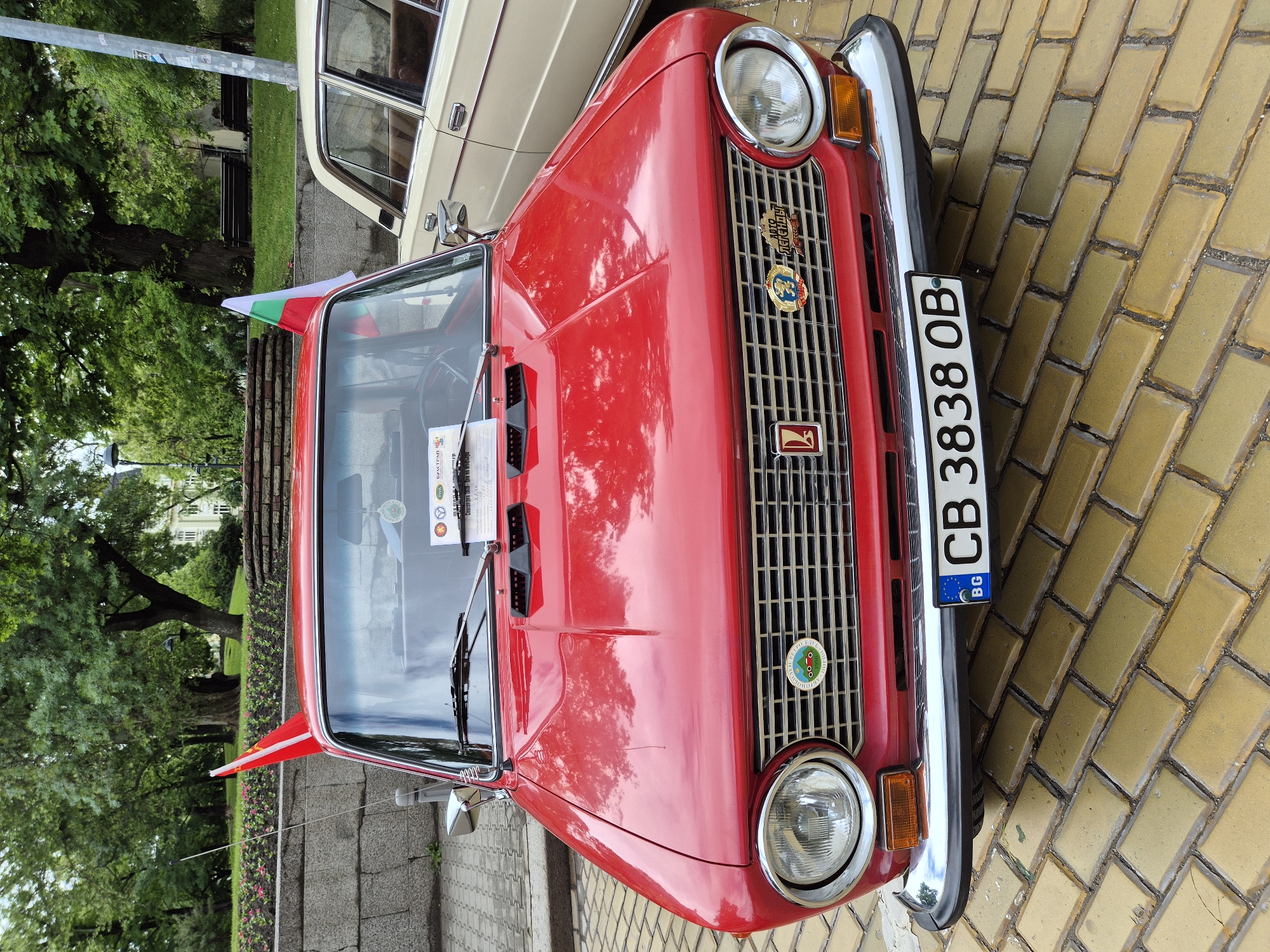 The image depicts a red Moskvich car, specifically an Aleko model, parked on a yellow brick-paved area. The car has a Bulgarian license plate reading "CB 3838 OB." A Bulgarian flag is visible on the left side of the car near the windshield. The background includes greenery with trees and a glimpse of another parked car, possibly a vintage model. The scene is likely located in a historical part of Sofia, Bulgaria, known for its characteristic yellow brick sidewalks. The image is shot from a slightly elevated angle, focusing on the front of the Moskvich.