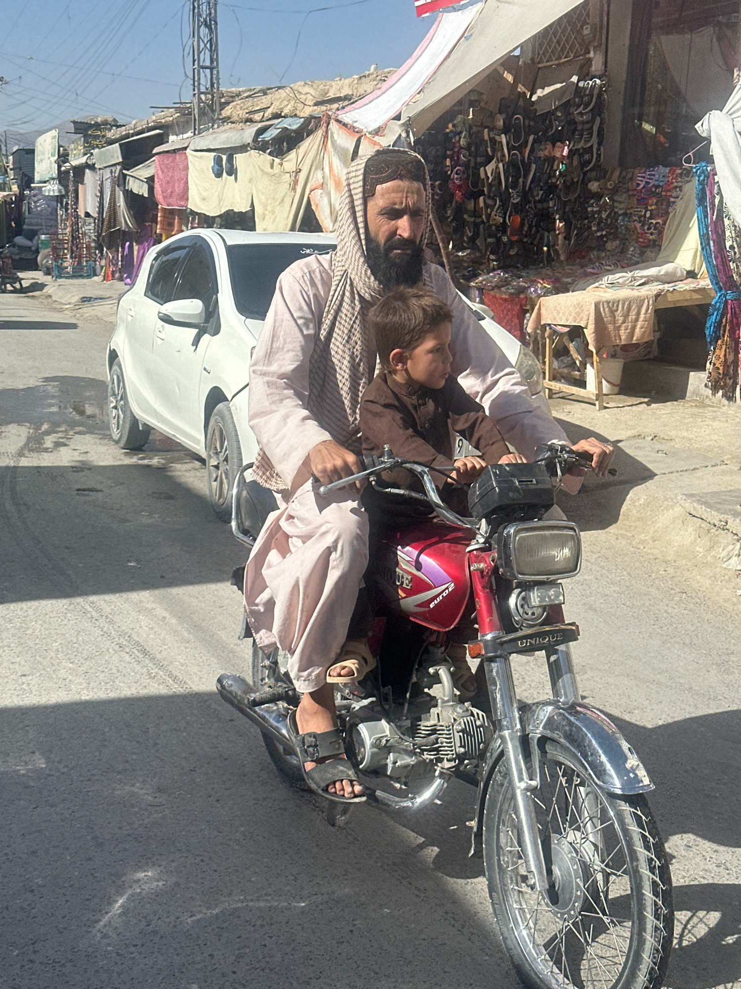 In Quetta City Tehsil, Pakistan, on a sunlit street with a cracked asphalt surface and tire tracks, a man and a child ride on a red motorcycle. The man, wearing a light pink shalwar kameez and a checkered scarf around his head, is the driver. He has a beard and is looking forward. The child, dressed in a brown outfit, sits in front of the man, also looking forward. The motorcycle has a black headlight, a silver fender, and the word "UNIQUE" is visible on the front. In the background, there are market stalls with various goods, including shoes and textiles, and a white car is parked nearby. The overall environment suggests a bustling street scene in a town.