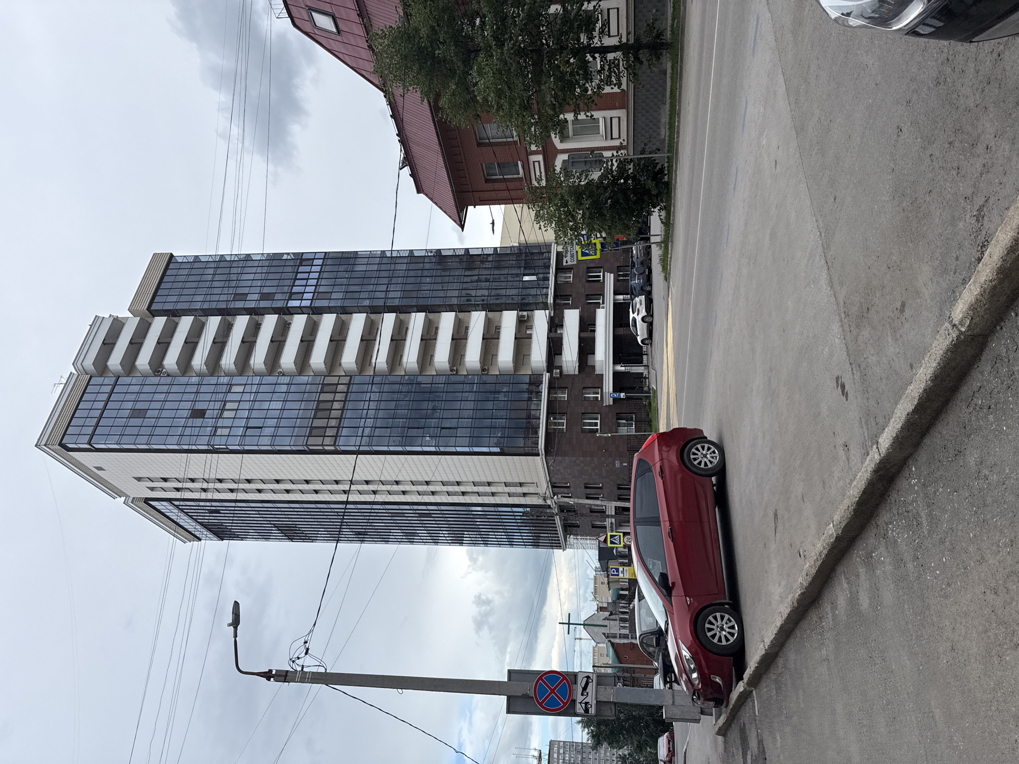 The image depicts a street scene in Perm, Russia, under an overcast sky. A modern, multi-story building with a glass facade and white balconies dominates the left side of the frame. To its right, a narrower, older brick building with red trim is visible. The street is paved with asphalt and bordered by a concrete curb.
Several cars are parked along the street. A bright red hatchback is parked parallel to the curb, occupying the foreground on the right. Behind it, other cars, including a white sedan, are parked in a row. The street has a yellow dividing line.
Traffic signs are present. One sign indicates parking, and another, a circular red sign with a blue cross and two diagonal lines, signifies a no stopping or no parking zone. A smaller sign below it shows a car being towed. A yellow pedestrian crossing sign is also visible.
A lamppost stands on the right side of the street, with electrical wires strung between poles. The overall atmosphere is that of a typical urban environment on a cloudy day. No people are clearly visible in the image.