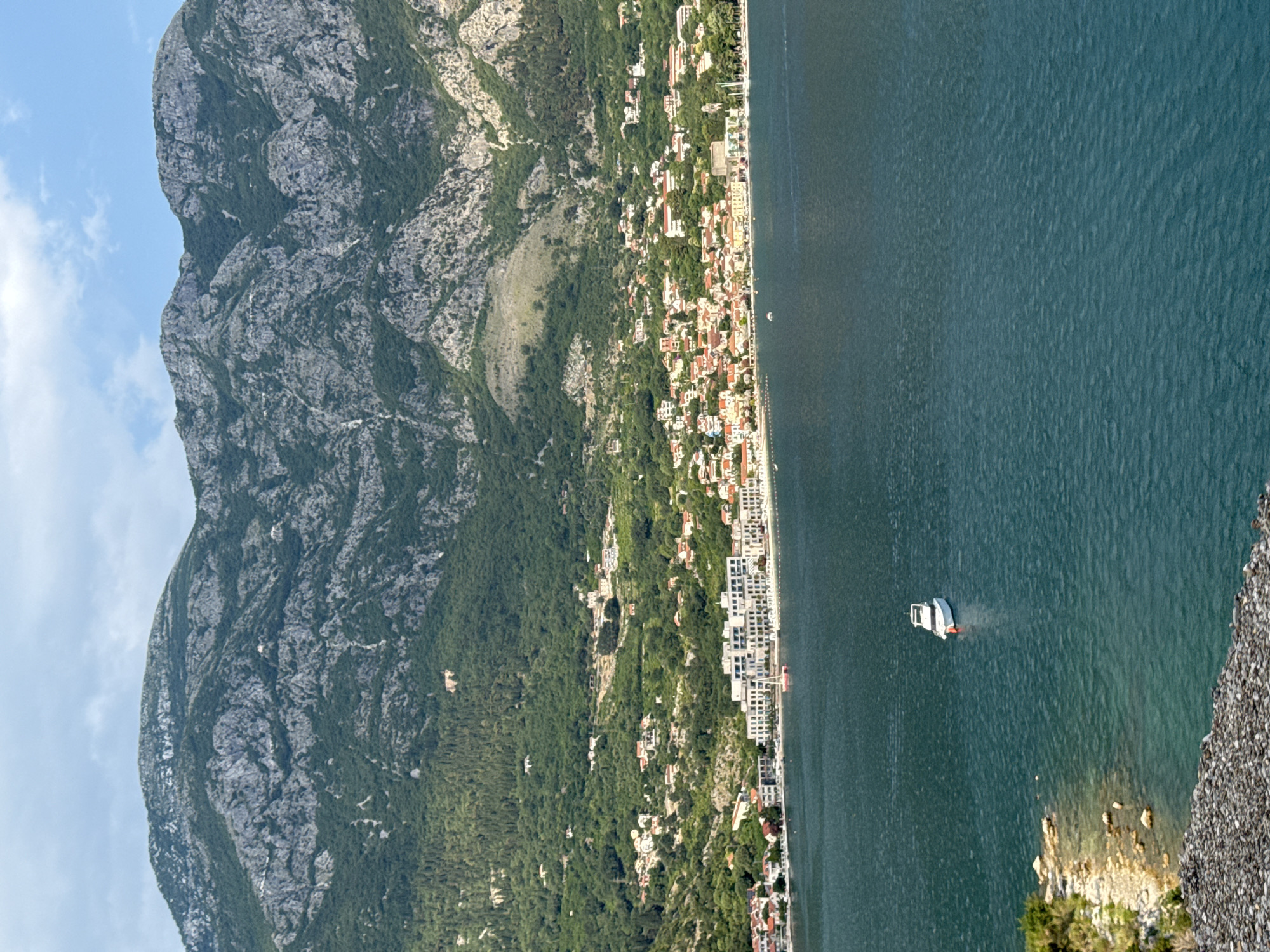 This is a landscape photograph of Risan, Montenegro, captured from an elevated perspective looking across the Bay of Kotor towards the town and its mountainous backdrop. The foreground is dominated by the teal-colored water of the bay, with a small white motorboat with a red bumper visible in the middle distance. To the left, a rocky shore with sparse vegetation lines the water's edge.
In the mid-ground, the town of Risan stretches along the coastline. Buildings with red-tiled roofs and white facades are clustered together, nestled into the hillside. A prominent modern white building complex with numerous balconies stands out along the waterfront. Further back, the slopes of the surrounding mountains are covered in dense green foliage, with rugged, rocky peaks rising above. The sky is a mix of light blue and scattered white clouds. The scene depicts a tranquil coastal town at the foot of imposing mountains.