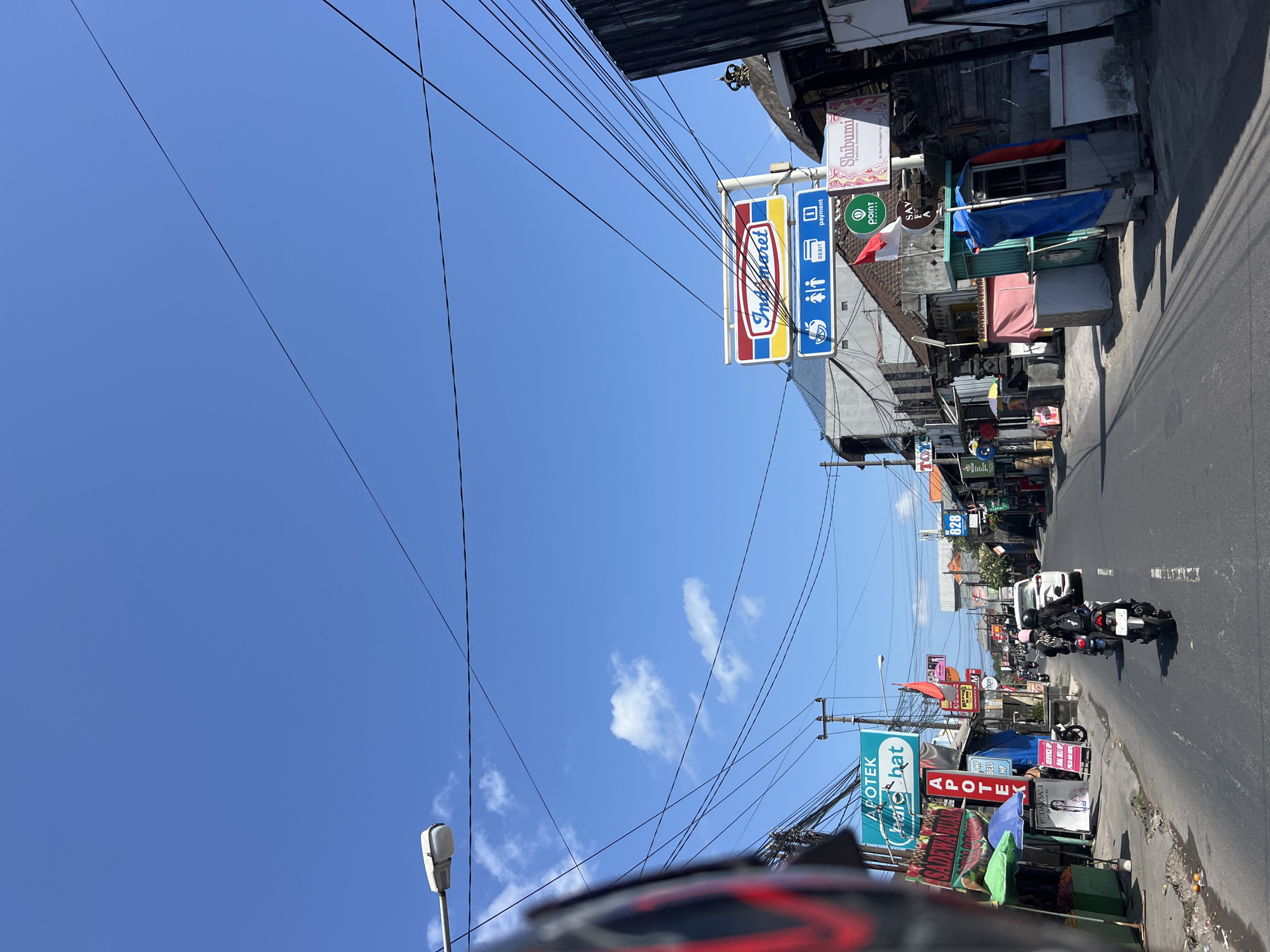 This is a street-level, forward-facing, eye-level shot on a sunny day in Denpasar, Indonesia. The scene depicts a busy street lined with shops and storefronts, with a clear blue sky overhead, dotted with a few wispy clouds. Electrical wires are prominently strung across the sky, supported by utility poles. The road is paved and appears to be a two-way street with white dashed lines marking the lanes.
On the left side of the road, there are various shops, including an "APOTEK Chaic Hat" (pharmacy) with a green awning, and other businesses displaying colorful signs. Some small stalls with umbrellas are set up along the sidewalk, and produce is visible in a green container. A portion of a vehicle's helmet, with red accents, is visible in the lower left corner, suggesting the photo was taken from a moving motorcycle or scooter.
On the right side of the road, there is a prominent "Indomaret" convenience store sign, with a smaller sign below indicating "DEBIT" and "payment." Further down the street, other shops and buildings are visible, some with traditional Balinese architectural elements like pitched roofs and ornate decorations.
Several people are present, primarily on motorcycles. Two individuals are riding a scooter in the middle of the road, and other motorcycles are seen further ahead. A white car is also on the road, indicating vehicular traffic. The overall impression is that of a typical commercial street in a Southeast Asian city, with a mix of modern businesses and local shops. The presence of multiple signs in Indonesian language, such as "APOTEK," "SADEWA RUAR," "WARUNG," and "POINT," confirms the location as Indonesia.