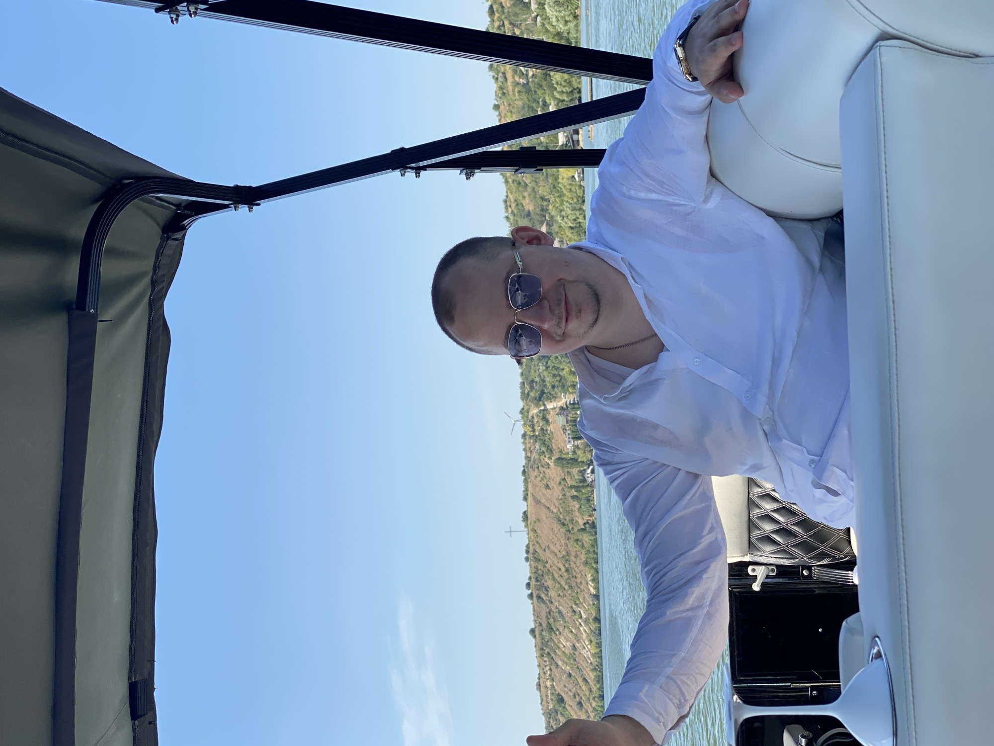 An undated, medium-shot photograph, taken from a low angle, captures a bald, fair-skinned man wearing a white collared shirt and sunglasses as he sits on a boat. He is looking directly at the camera, with his left arm resting on a white upholstered seat and his right arm extended with his thumb raised. The boat's interior, featuring black and white upholstery and a white cup holder, is visible in the foreground. The background depicts a body of water, a green, hilly landscape, and a clear blue sky. The man is positioned in the city of Molovata, Moldova.