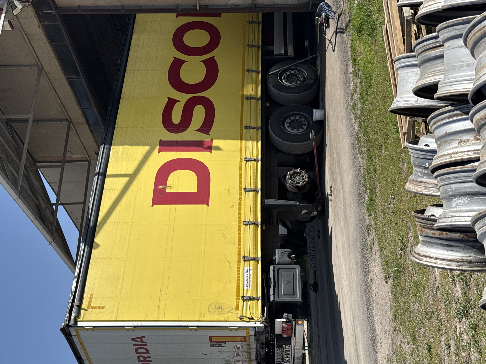 A large yellow truck trailer with "DISCO" emblazoned in prominent red letters on its side is depicted, parked alongside an industrial building or loading structure. The image, taken in Busmantsi, Bulgaria, shows the trailer oriented horizontally, with its bright yellow tarpaulin side facing the viewer. Underneath the trailer, multiple truck tires are visible, as well as parts of the chassis and suspension. A coiled air hose hangs from the side of the truck near the cab area.
To the right of the trailer, a stack of numerous metal wheel rims, some appearing weathered or used, are neatly piled on the ground next to a grassy patch. Further right, a dirt and gravel path or road runs parallel to the trailer. On the left side, the metallic framework of a roof or canopy extends over the truck, casting shadows from the bright daylight. The sky is clear and blue, indicating sunny weather.
At the rear of the truck (bottom left of the image), a portion of another white trailer is visible, with partial text "ORDIA" and a license plate holder bearing the name "SCHMITZ". A smaller label on the yellow trailer also shows "SCHMITZ CARGOBULL". The scene suggests an outdoor industrial setting, possibly a transport depot, warehouse, or vehicle service center, where the truck is either parked or awaiting loading/unloading.