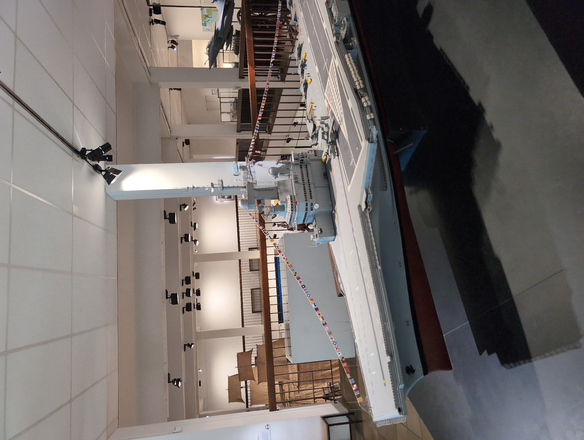 The image depicts a model of an aircraft carrier on display within a building. The carrier model is predominantly grey, with a deck marked with white lines and various miniature aircraft. The ship's hull has a red section. Hanging nearby is a decorative string of nautical signal flags. In the background are architectural features including white walls, support pillars, and rows of lights, as well as a model sailing ship behind a railing. It is a museum of the French Navy, located in Toulon, France.