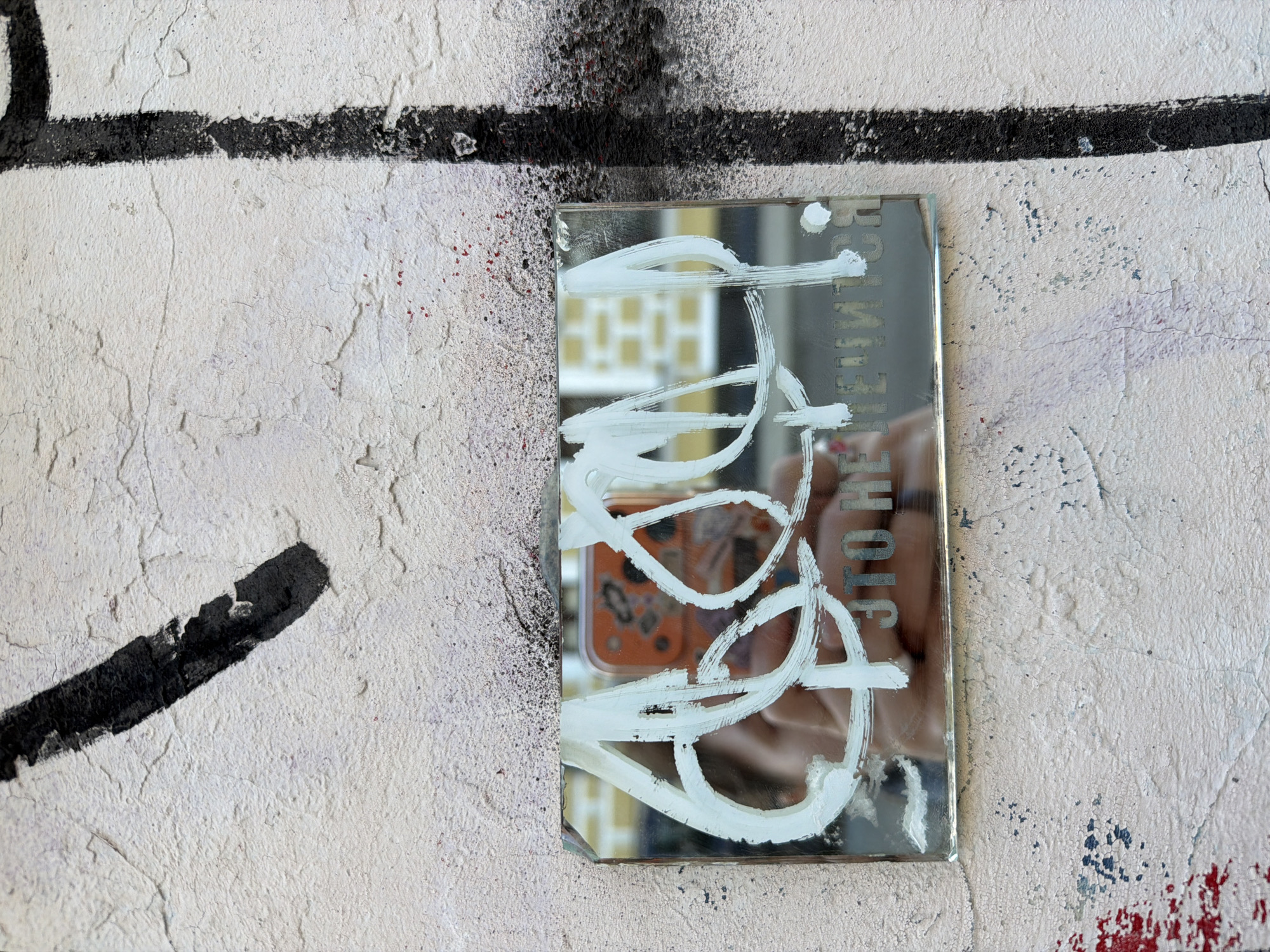 An urban scene features a rectangular mirror placed against a textured, off-white wall in Moscow, Russia. The wall's uneven surface shows cracks and wear, adorned with black spray paint, including a thick horizontal line across the upper section and a shorter diagonal stroke in the lower left. Splashes of red and blue/grey paint are also visible, particularly on the right side.
The mirror's reflective surface is largely obscured by bold, white, brush-stroked graffiti, forming abstract, sweeping patterns. Reflected in the mirror is a person holding a smartphone with an orange or reddish-brown case, indicating the act of photography. The person's hand and a portion of their chin/jawline are visible. Beyond the person, an architectural background with distinct square or rectangular window patterns can be seen, suggesting a building in an urban street setting.
Etched or engraved into the mirror, beneath the white paint, is text in Russian Cyrillic. Due to the reflection, the letters appear reversed, but they spell "ЭТО НЕ ИЛЛЮЗИЯ" (ETO NE ILLYUZIYA), which translates to "THIS IS NOT AN ILLUSION." The text is capitalized. The scene is illuminated by bright, even light, suggesting it is daytime. The mirror, with its painted message and self-referential reflection, acts as a piece of street art integrated into the gritty urban environment.