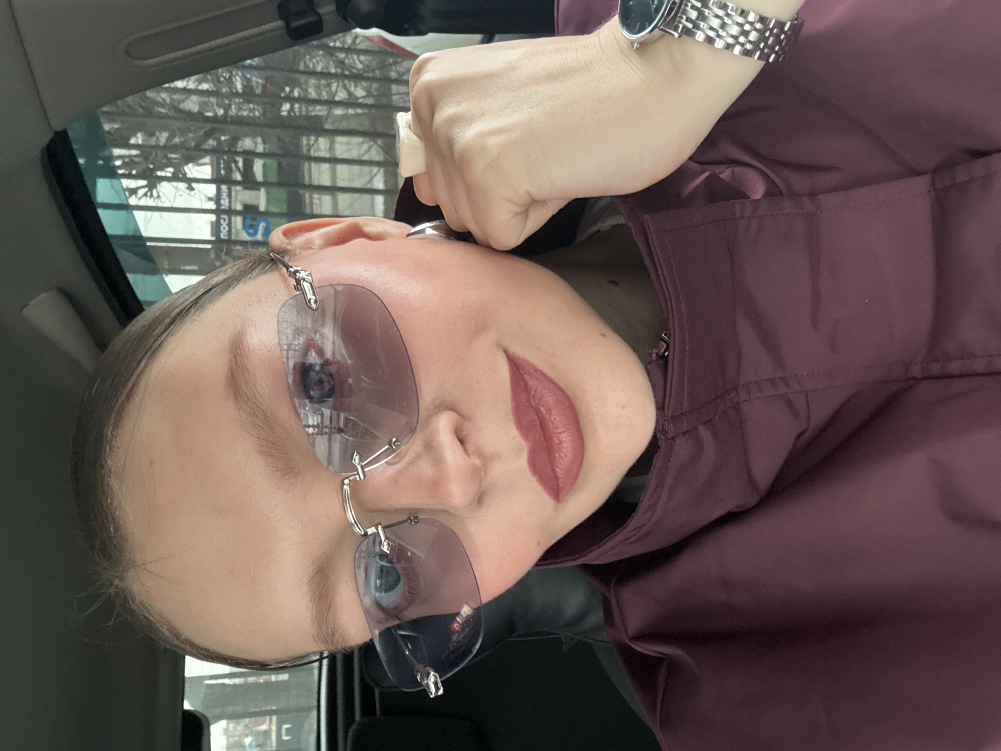 A woman is positioned inside a vehicle, likely a car, as suggested by the interior elements. She is wearing rimless sunglasses with a pale tint. She has fair skin, light eyebrows, and is wearing a dark matte lipstick. The woman is wearing a metal watch on her left wrist. She is wearing a burgundy jacket or coat. Her left hand is raised and clenched into a fist. The reflection in the sunglasses and the view out the vehicle's windows show an urban area in Izhevsk, Russia, with bare trees, and street signs in Russian.