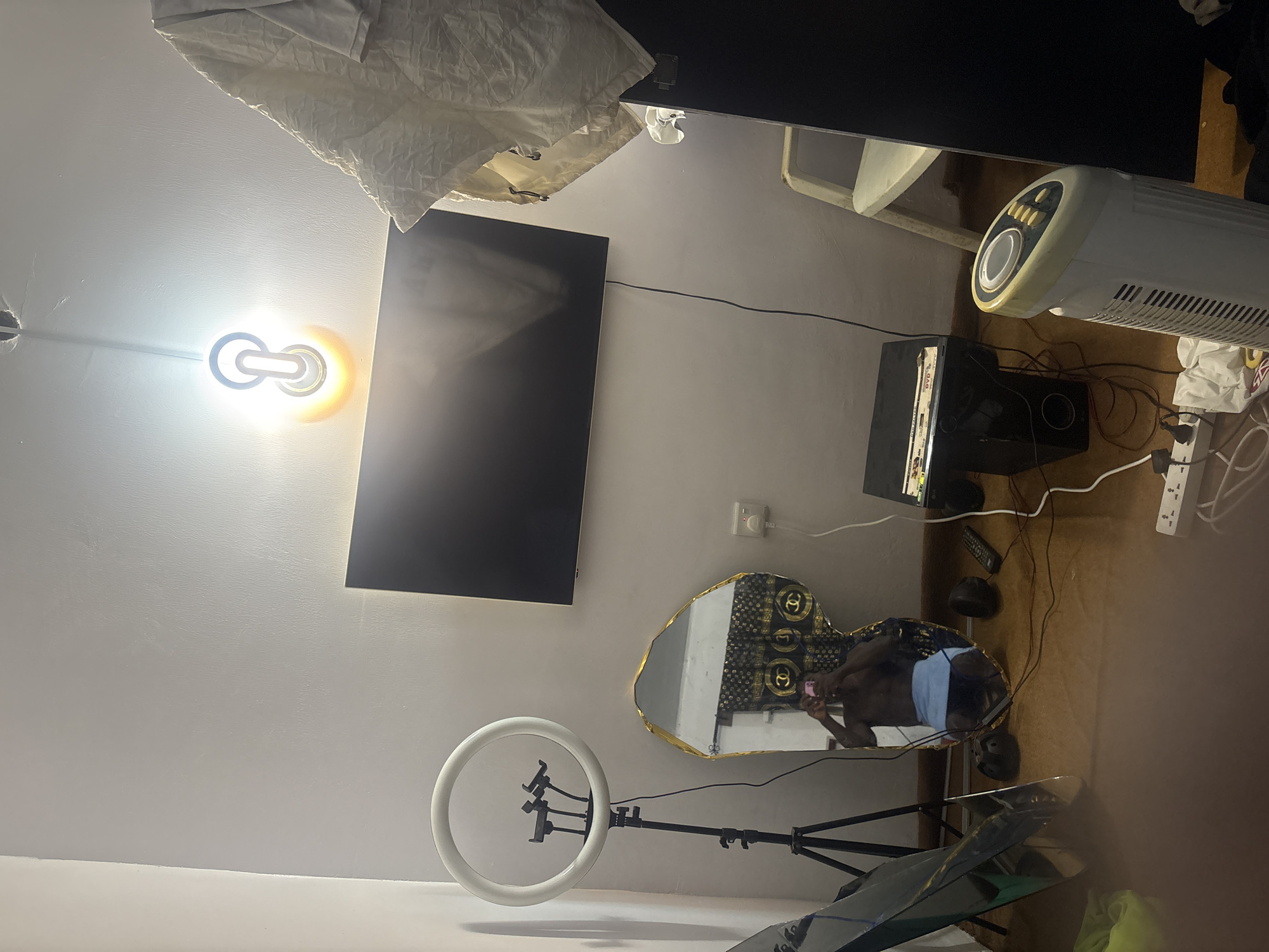 A room in Bukuru, Nigeria contains a flat-screen TV mounted on a white wall, above a heart-shaped mirror in which a person is visible, taking a selfie. A ring light on a stand is positioned near the mirror. A white fan heater is on the right. Other items include a speaker, and miscellaneous cables and items on the floor. A white sheet hangs from a shelf on the upper left. A light fixture is on the wall near the TV.