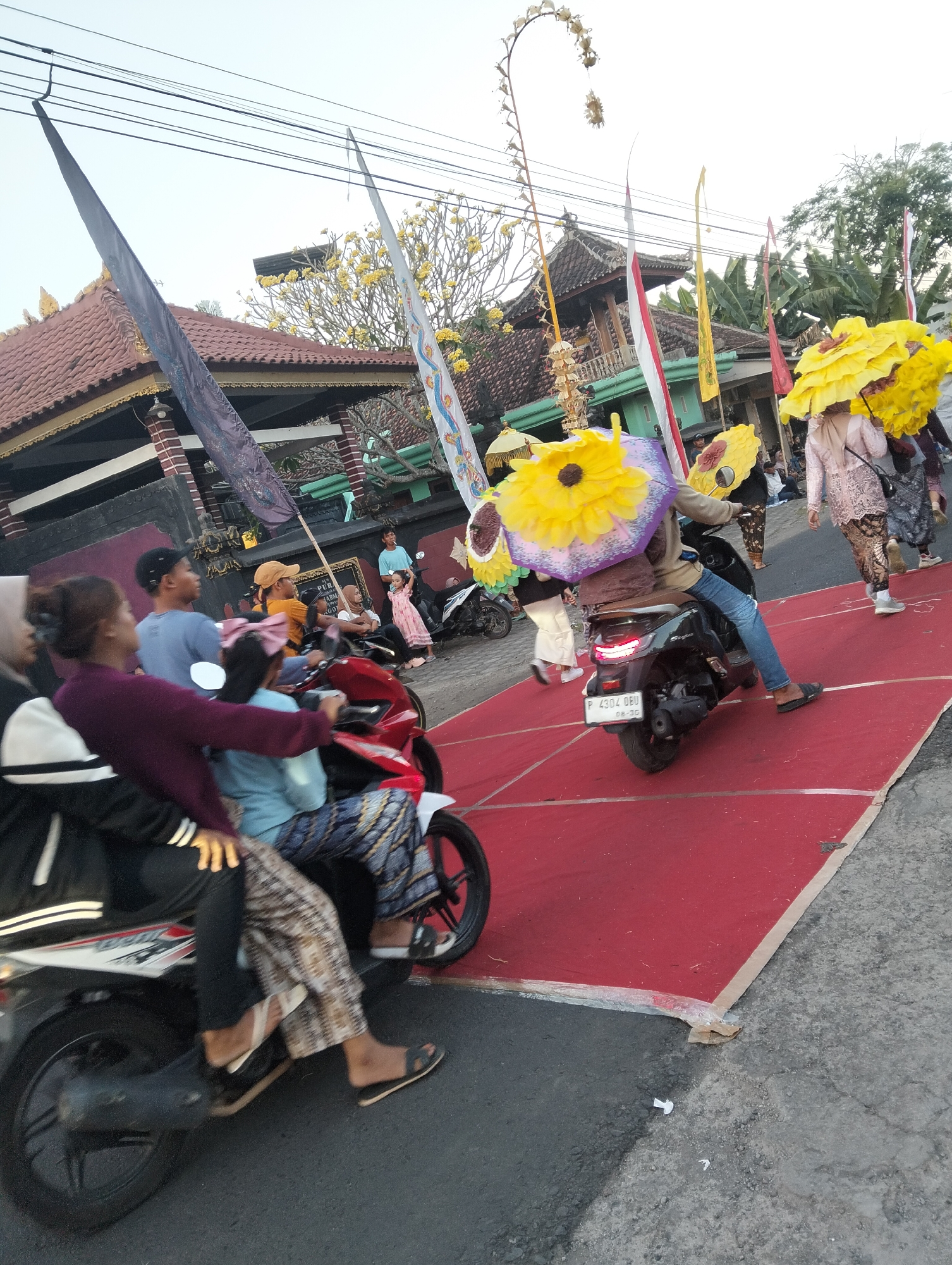 In Sumbersewu, Indonesia, a procession is underway on a street adorned with a red carpet. On the left, two individuals are riding a red scooter, the passenger wearing a maroon long-sleeved shirt and the driver a blue shirt. Behind them, on a black scooter, a person wearing blue jeans and a grey shirt navigates the street, holding a large yellow sun-shaped prop. Further back, other participants are seen, some holding similar yellow props, dressed in traditional attire including a light pink lace top and a dark patterned skirt. Several flags are visible, including red and white Indonesian flags and yellow flags, adding a festive atmosphere to the scene. Traditional Indonesian buildings with tiled roofs line the street, and trees with yellow flowers bloom in the background. The sky is overcast, indicating a late afternoon or early morning light. The street is paved, with some debris scattered on the ground near the red carpet.