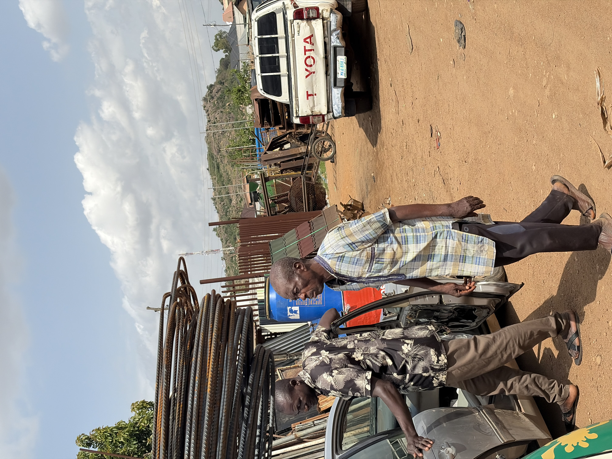 The image depicts an outdoor commercial or work scene, likely in Gura Top, Nigeria, characterized by materials such as rebar and scrap. Two adult men are visible in the foreground, engaged in activity around a car.
On the right, one man is seen from the side, wearing a blue, yellow, and white plaid shirt, dark trousers, and sandals. He stands upright, looking towards the car. On the left, another man, dressed in a dark shirt with a light floral pattern and brown trousers, also wearing sandals, is leaning into the open driver's side door of a grey sedan. They appear to be interacting with the vehicle, possibly in the process of loading or unloading.
Dominating the immediate left foreground is a large, tightly coiled stack of rusted rebar. Behind the men and the car, there are several industrial barrels, including a blue one and a red one. Further back, a white Toyota Hilux pickup truck is parked, clearly displaying "TOYOTA" in red letters on its tailgate and "HILUX" vertically on the left side. Its license plate, "MKA-4HA1", is visible. Nearby, a two-wheeled cart and various pieces of scrap metal are scattered on the reddish-brown dirt ground, with corrugated metal sheets forming a barrier or structure behind them.
The scene unfolds under a bright, clear sky with scattered white clouds, indicating it is daytime. In the background, rocky hills with some vegetation rise, suggesting a semi-urban or rural environment. The overall impression is one of everyday work or commerce.