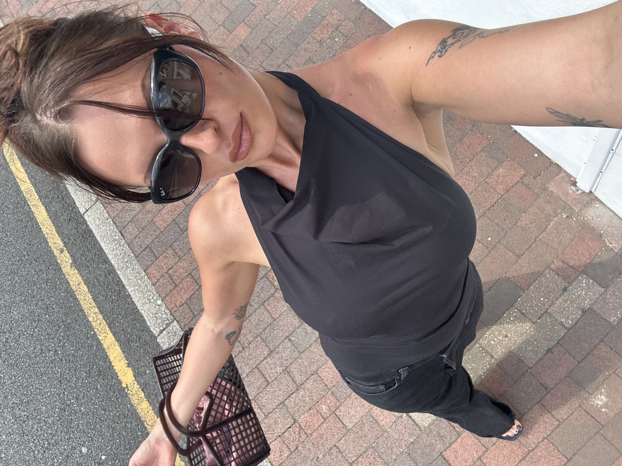 A woman stands on a brick sidewalk adjacent to a street with a yellow line. She is wearing a black halter top and black jeans, and is carrying a black shopping basket. She is also wearing black sunglasses. The woman has her hair pulled up in a bun and has tattoos on her arm. The overall scene suggests a casual outing in a town setting.