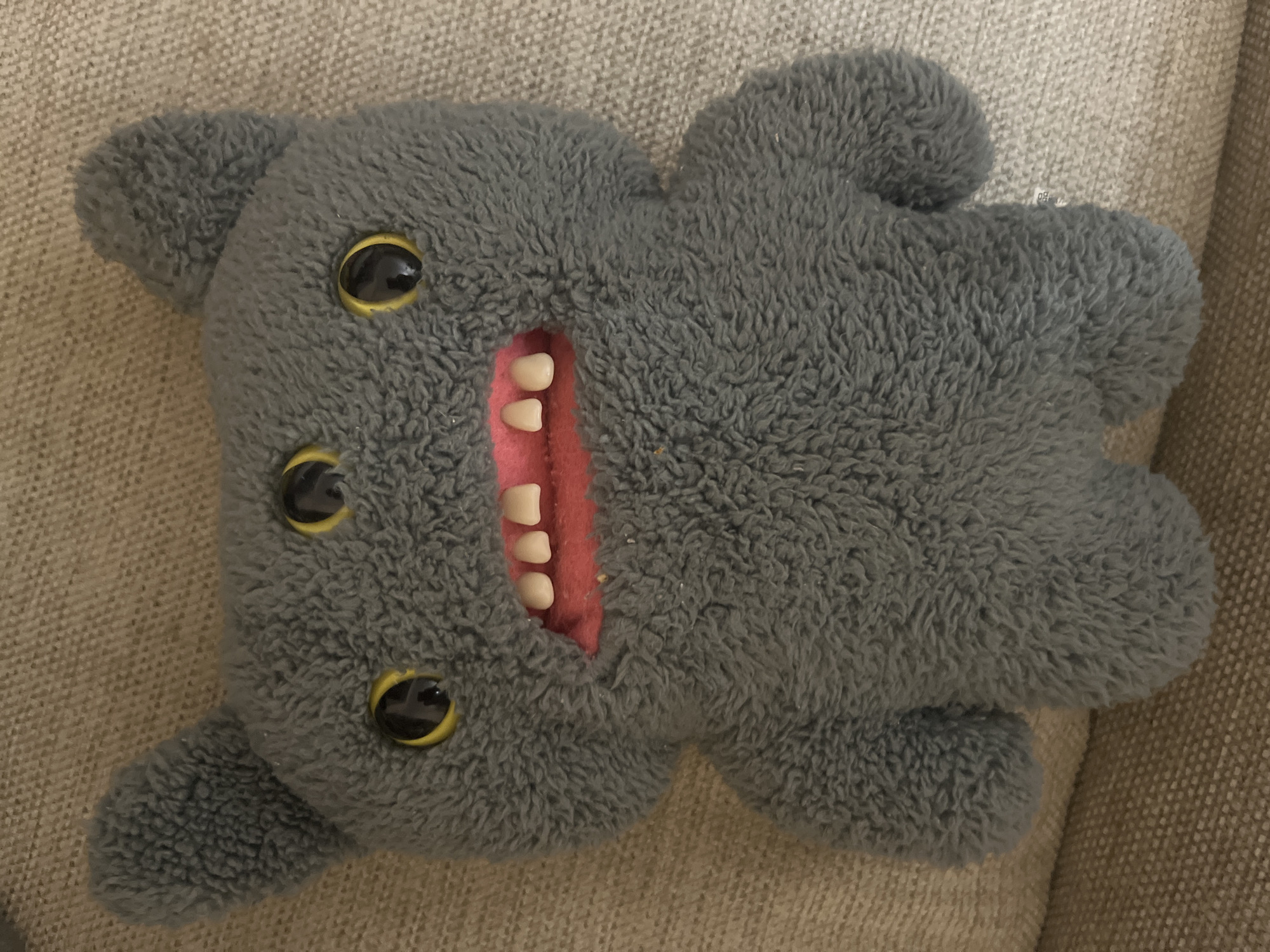 A gray, fuzzy plush toy with three yellow-eyed black eyes and large, pink lips with four visible teeth is shown from the chest up, resting on a textured beige surface. The toy has two small, rounded ears on top of its head. The lighting is neutral, and the image appears to be a close-up shot taken indoors. There is no indication of the city of Aktau, Kazakhstan in the image or its contents.
