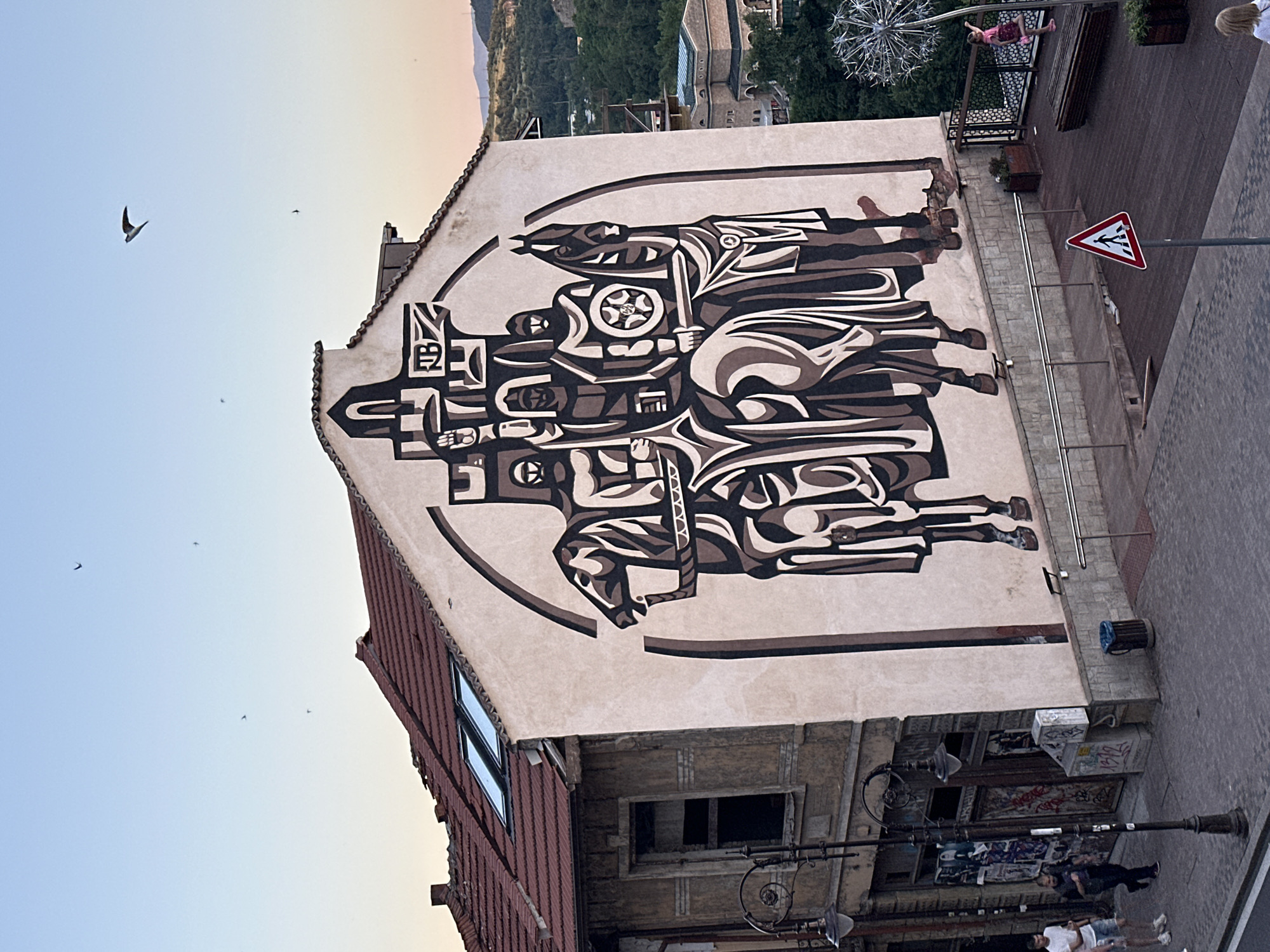The image captures a mural on the side of a building in Veliko Tarnovo, Bulgaria. The mural depicts a stylized, monochromatic scene of figures on horseback, rendered in shades of brown and beige with black outlines. The figures are clad in historical-looking attire, with helmets and shields visible. The mural covers a significant portion of the building's facade, arching towards the roof.
The building itself appears to be old, with weathered walls and a terracotta tiled roof featuring dormer windows. Adjacent to the building with the mural, another structure stands, showing signs of disrepair with a broken window. A street lamp with an ornate design stands on the sidewalk in front of this older building.
Below the mural and the buildings, a paved walkway and a wooden deck area are visible. Several people are present in the scene. Two men are walking on the sidewalk near the street lamp. Further along the deck, a young girl in a pink dress is interacting with a large, metallic dandelion sculpture. A bench is situated near the sculpture. A pedestrian crossing sign is positioned on the walkway, indicating a street or pathway. In the background, lush green hills dotted with trees and structures can be seen, suggesting the hilly terrain of Veliko Tarnovo. Several birds are flying in the clear sky above the buildings. The lighting suggests it is late afternoon or early evening.