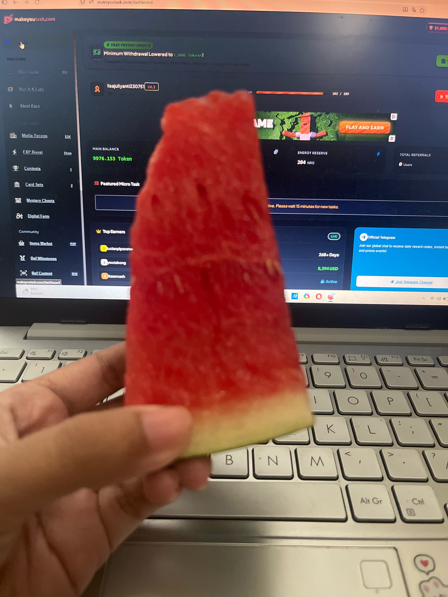 A hand holds a slice of watermelon in front of a laptop screen displaying the website makeyoutask.com. The website shows various user statistics and tasks. The laptop is open on a table. The overall scene suggests a person multitasking, likely working or browsing online while enjoying a snack. There is no information in the image that can provide details about the location in the city of Meliau Hulu, Indonesia.