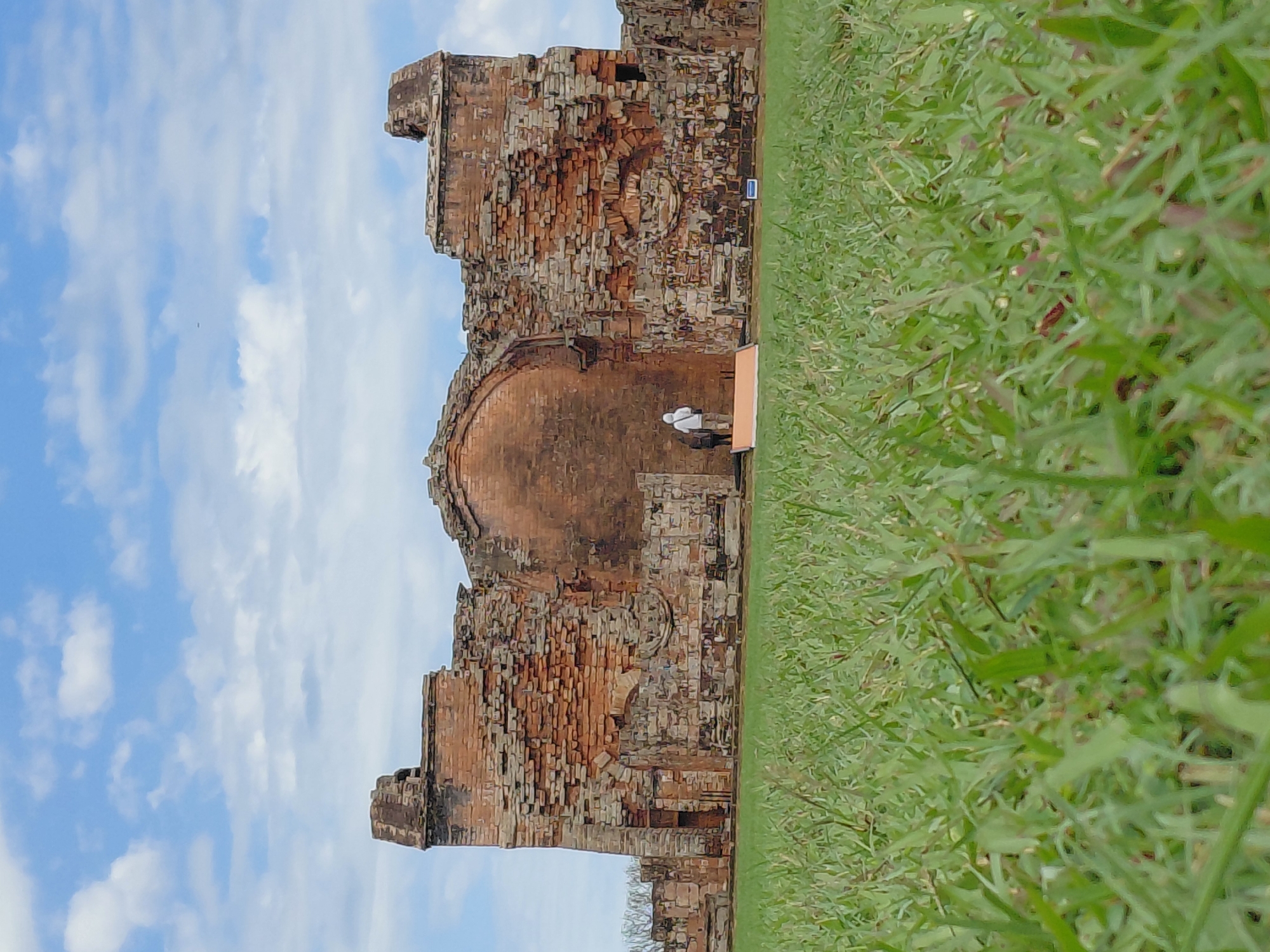 The image captures a view of the ruins of the Santísima Trinidad de Paraná Jesuit mission, located in Trinidad, Paraguay. The foreground is dominated by lush green grass, viewed from a low angle, giving a sense of depth and scale. In the mid-ground, the remains of the mission stand, constructed from weathered red brick and stone. The structure is partially intact, with crumbling walls, arched doorways, and decorative stonework. Two individuals are visible standing near the center of the ruins, appearing to observe the site. The background consists of a bright blue sky with scattered white clouds, suggesting a clear day. The overall scene evokes a sense of historical significance and natural beauty.