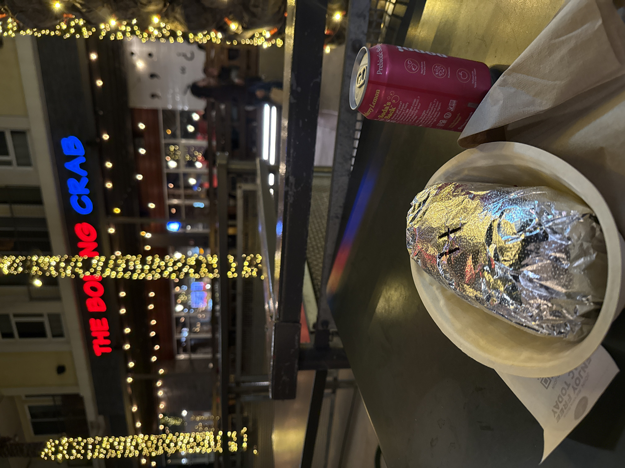The image shows a meal on a table outdoors at "The Boiling Crab" restaurant in Burbank, California, United States of America. The meal consists of a burrito wrapped in foil in a bowl, a napkin, and a can of "Soda's Back" with berry lemon flavor. The restaurant’s sign is visible in the background, along with Christmas lights. People are walking in the background. The image appears to have been taken in the evening.