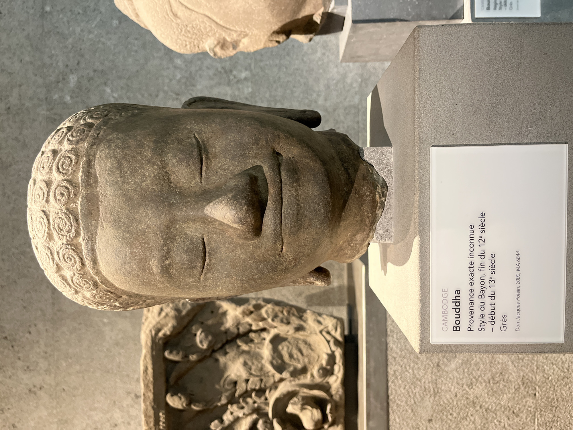 The image shows a stone sculpture of a Buddha's head displayed on a grey pedestal. The sculpture is in the center of the frame, facing forward with closed eyes and a gentle smile. The Buddha's hair is depicted as a series of tightly coiled curls. To the left of the Buddha's head, another stone artifact, possibly a relief carving, is visible. To the right, another Buddha's head sculpture is partially visible, also on a pedestal. A white plaque with text is attached to the pedestal in front of the main sculpture. The text on the plaque reads: "CAMBODGE", "Buddha", "Provenenance exacte inconnue", "Style du Bayon, fin du 12e siècle - début du 13e siècle", "Grès", and "Don Jacques Polain, 2000, MA 6844". The background is a textured grey wall. This appears to be an exhibit in a museum. Given the context of museum exhibits and the specific item labels often found in such institutions, this image is likely from the Musée Guimet in Paris, France, which houses Asian art.