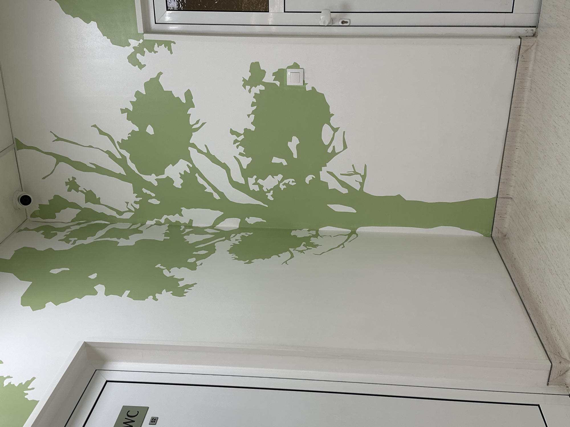 The image shows a section of a restroom wall in a building, possibly in Sofia, Bulgaria. The wall is decorated with a light green tree mural painted on a white background. Visible architectural features include a door frame at the bottom and a window frame near the top. A small surveillance camera is mounted on the wall near the top left corner. A light switch is visible above the tree mural. The wall has a slightly textured surface. The image appears to be taken from a low angle, looking upwards.