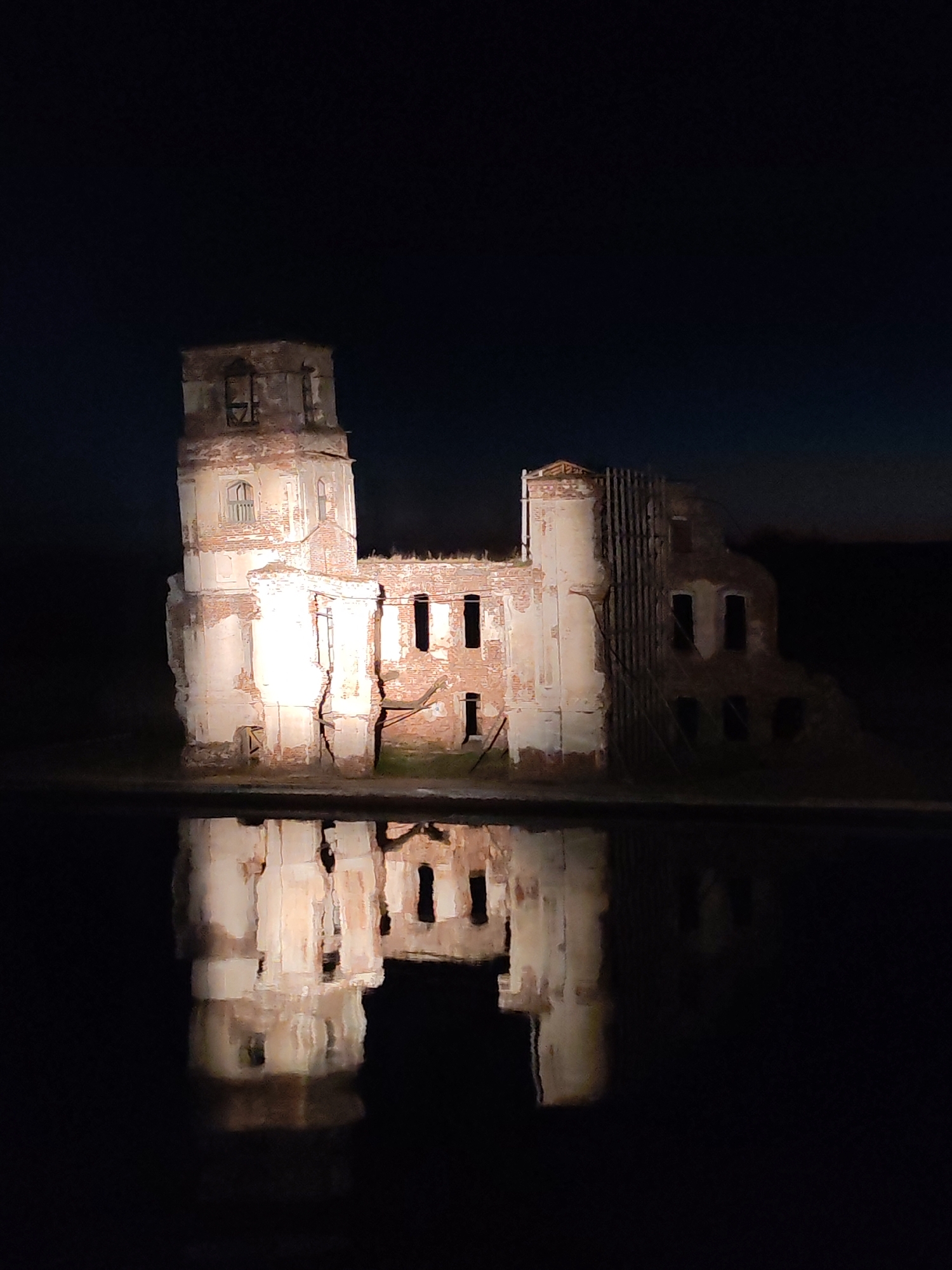An eye-level long shot depicts the ruined Church of the Nativity in Krokhino, Russia, at night. The pale exterior walls contrast sharply against the nearly black sky and dark water reflection below. The church is partially destroyed with gaping window holes and crumbling structure. The building is illuminated, causing the walls to glow and emphasizing the damage. A dark reflective pool surrounds the church, mirroring its structure.