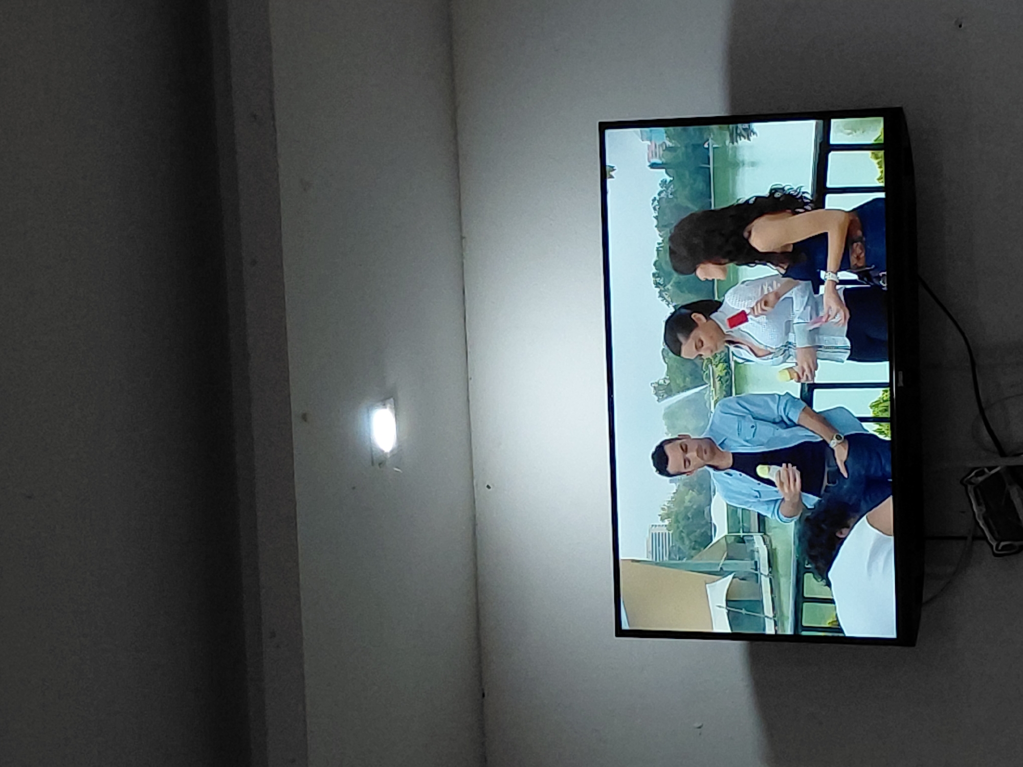 The image captures an indoor scene, primarily focusing on a television screen mounted on a plain, light-colored wall. The room appears to be sparsely furnished, with a bright, glowing light fixture attached to the wall on the left side of the frame, suggesting it is illuminated. Dark cables are visible extending from behind the television, leading down to a small, dark wall-mounted device or plug below the screen. The image itself is rotated 90 degrees counter-clockwise.
The television displays an outdoor scene, likely from a show or commercial, set during the daytime under clear weather. The setting appears to be a park or waterfront area, featuring a body of water (possibly a lake or pond) surrounded by green trees and distant buildings in the background. A dark railing or fence separates the foreground characters from the water.
At least four individuals are visible on the TV screen. In the upper half, a woman with long, dark curly hair wearing a dark top stands beside another woman with dark hair dressed in a light-colored, possibly white or pale blue, collared shirt. The woman in the light shirt holds a red and a yellow object, which resemble popsicles or small bottles. Below them, a man wearing a light blue button-up shirt and dark pants holds a white bottle. Partially visible in the bottom foreground of the TV screen is another person with dark hair, wearing a white t-shirt, who appears to be bending over. The general atmosphere of the TV content seems casual and relaxed. There is no visible text on the screen or in the surrounding room. The location context for the room is Santa Ana, Venezuela.