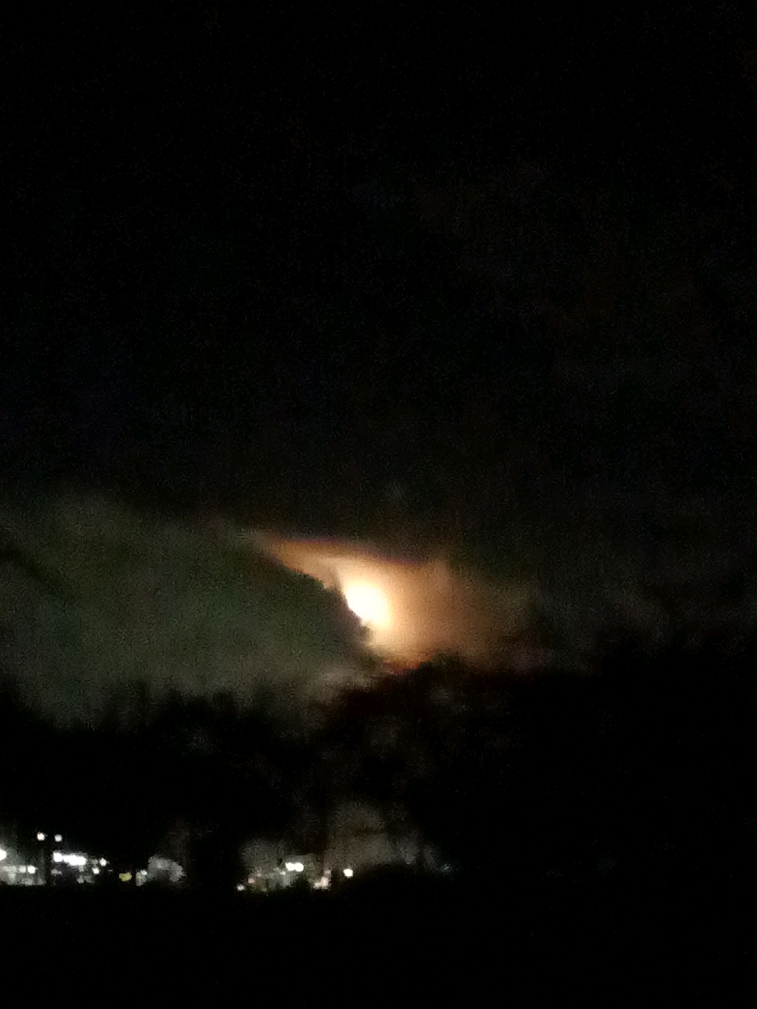 A dark, grainy, vertical outdoor photograph captures a night scene in Las Guevaras, Venezuela. The upper portion of the image is dominated by black sky, with a bright, nebulous orange and white glow emanating from a cloud formation in the center. This light appears to be the moon obscured by clouds, casting an ethereal illumination. Below this celestial phenomenon, the dark silhouettes of trees and foliage are visible. In the lower section of the frame, a distant line of blurred, out-of-focus lights suggests the presence of a town or city, with scattered white and yellow points of light indicating buildings or streetlights. The overall atmosphere is dim and somewhat mysterious, with the glow from the sky providing the primary source of light.