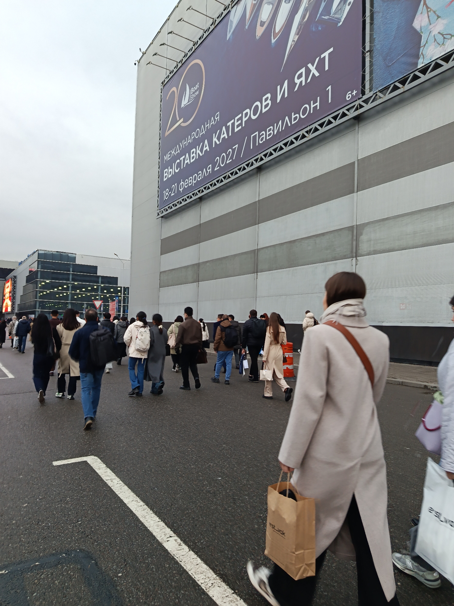 The image captures a street scene in Khimki, Russia, with a crowd of people walking along a pedestrian crossing. In the background, a large building displays a prominent billboard advertising the "Boat Show," scheduled for February 18-21, 2027, in Pavilion 1. The billboard includes the "6+" age restriction.
The crowd is diverse, with people wearing a variety of coats, jackets, and trousers. Some individuals carry bags. The person closest to the camera is wearing a light-colored coat and carrying a brown paper bag.
The sky is overcast, suggesting a cloudy day. The building has a horizontal striped pattern, and the overall scene conveys an urban atmosphere. The street itself is marked with pedestrian crossing stripes.