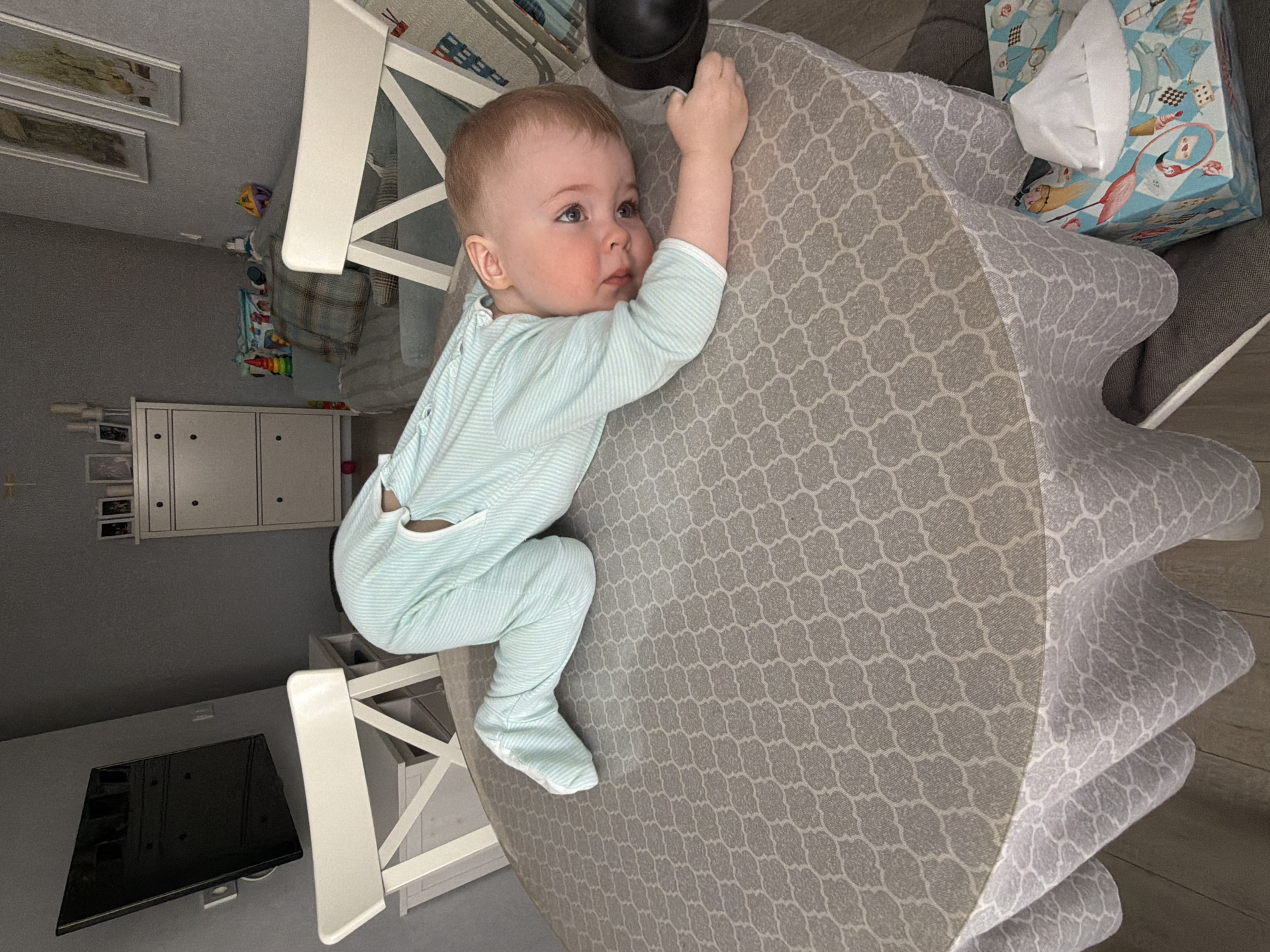 An infant is positioned face-down on a round table covered with a grey quatrefoil-patterned tablecloth within a domestic setting in Votkinsk, Russia. The baby, dressed in a light blue and white striped romper with attached footies, is propped on its forearms, with its head turned right, looking towards the viewer. Its cheeks are flushed, and its right hand holds a matte black, cup-shaped object. Two white wooden chairs with X-backrests are partially visible near the table. In the background to the left, a wall-mounted black flat-screen television is visible on a grey wall. To the right, a white two-drawer dresser topped with framed photos and candles stands against another grey wall, which also displays two framed pictures. A light blue sofa with plaid cushions and several children's toys (stacking rings, blocks) is visible further to the right. A decorative tissue box rests on the floor or a low surface beneath the table, to the right. The flooring is light-colored wood.
