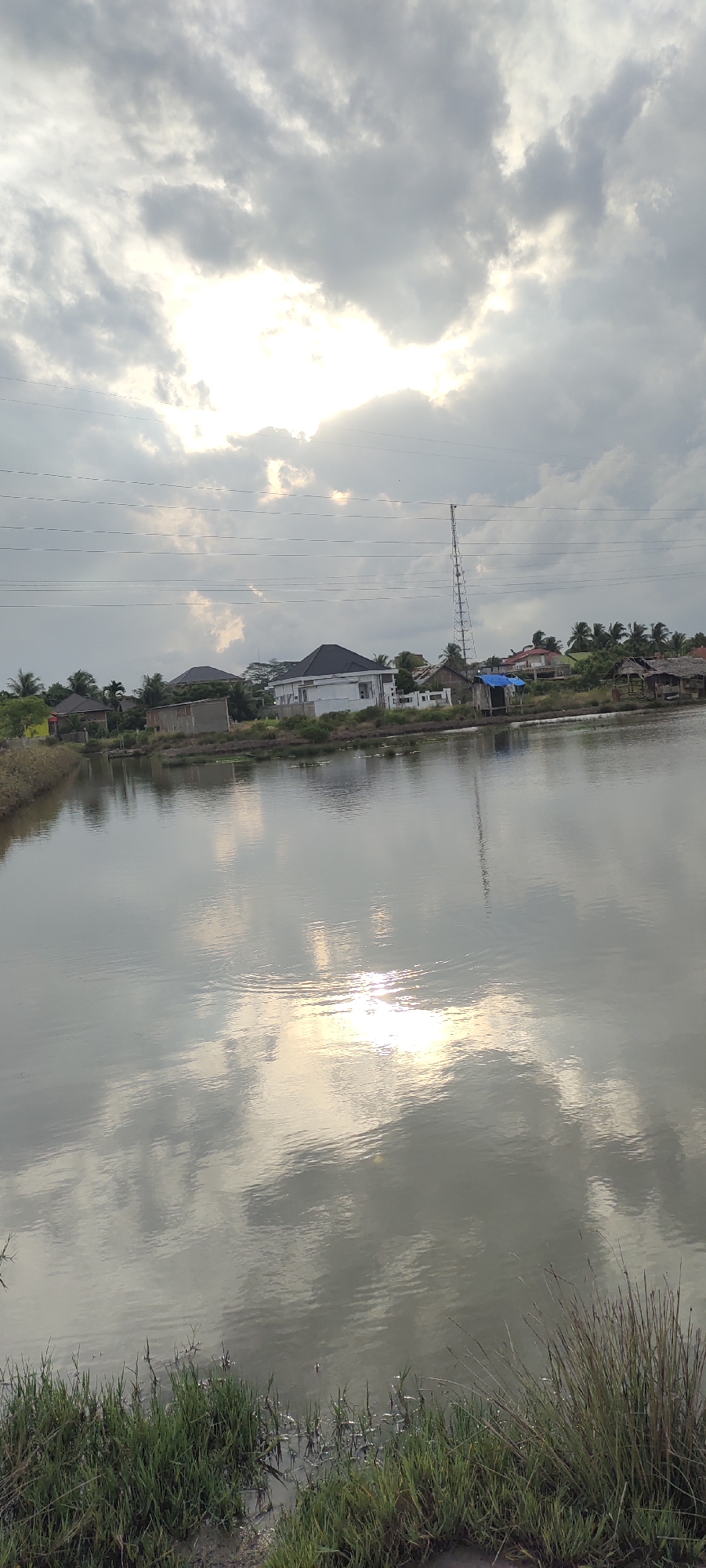 The image depicts a scene near a body of water, possibly a pond or lake, in Lhokseumawe, Indonesia. The sky is overcast with scattered clouds, and the sun is visible through a break in the clouds, casting a bright reflection on the water's surface. In the background, a row of houses and buildings can be seen along the bank, with some trees interspersed. A tall communication tower stands prominently among the structures. The foreground features the edge of the water with tall grass growing on the bank. The water is calm, with ripples indicating slight movement. The overall atmosphere is serene and natural, with a touch of human presence in the form of the buildings and the tower.